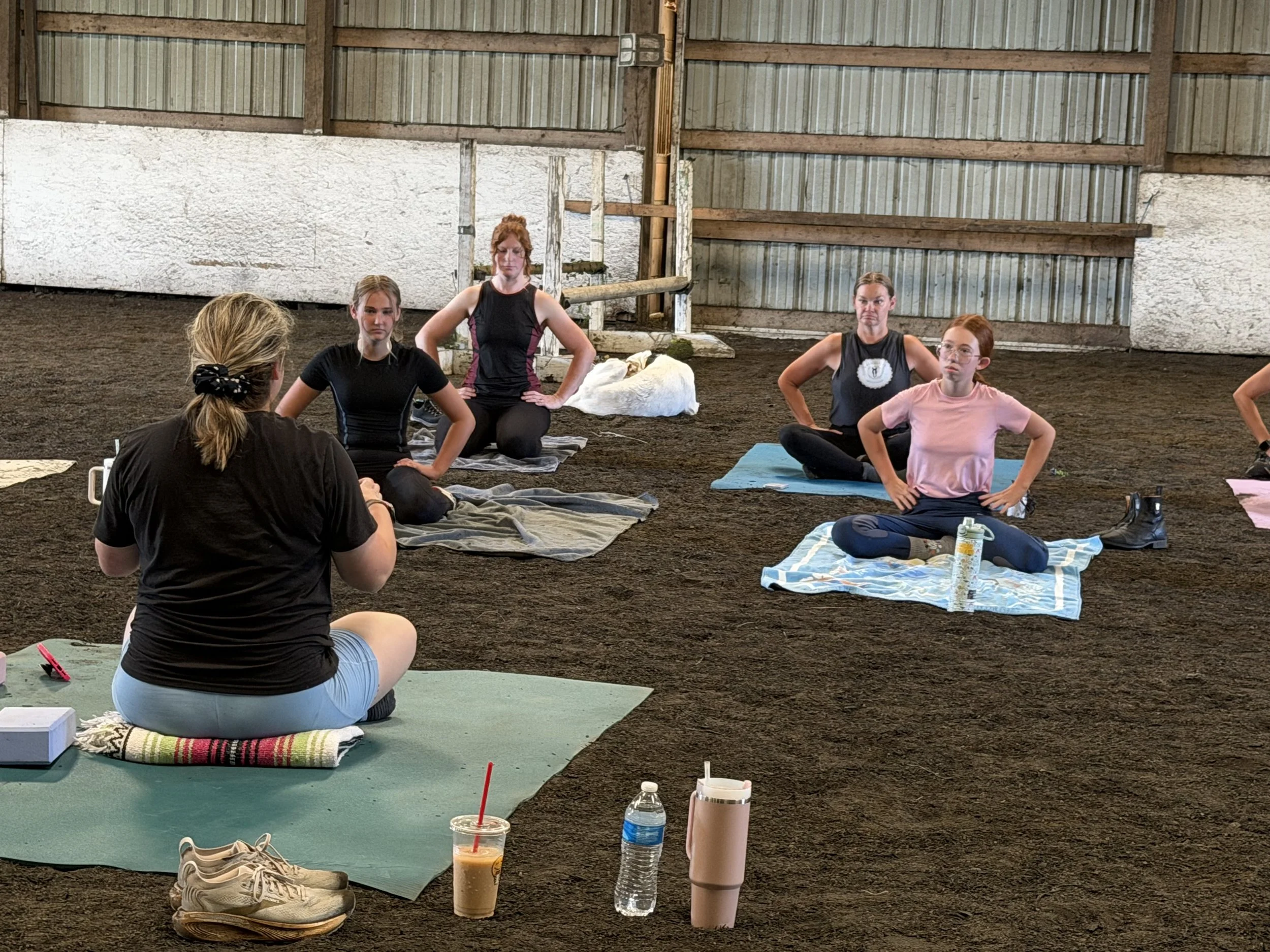 Women participating in a yoga or meditation class inside a rustic barn with dirt floor, seated on yoga mats and towels, with water bottles and personal items nearby.