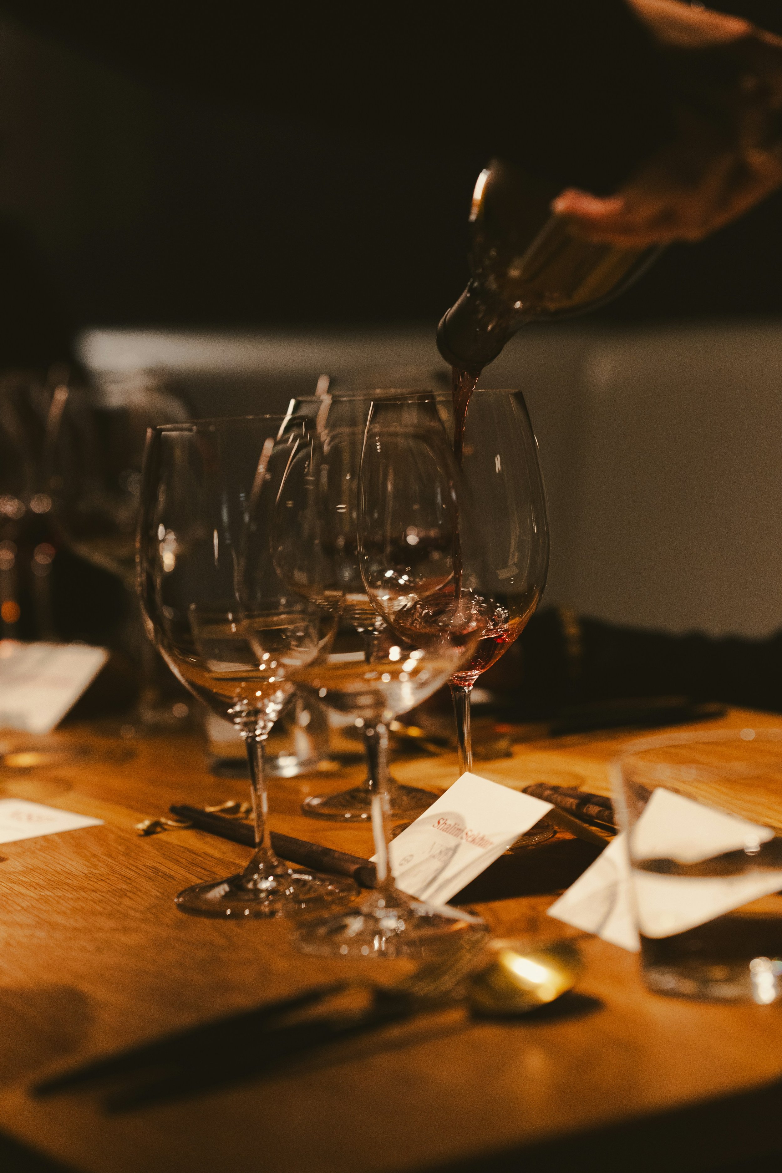 Red wine being poured into a glass on a wooden table with other empty glasses and place cards.