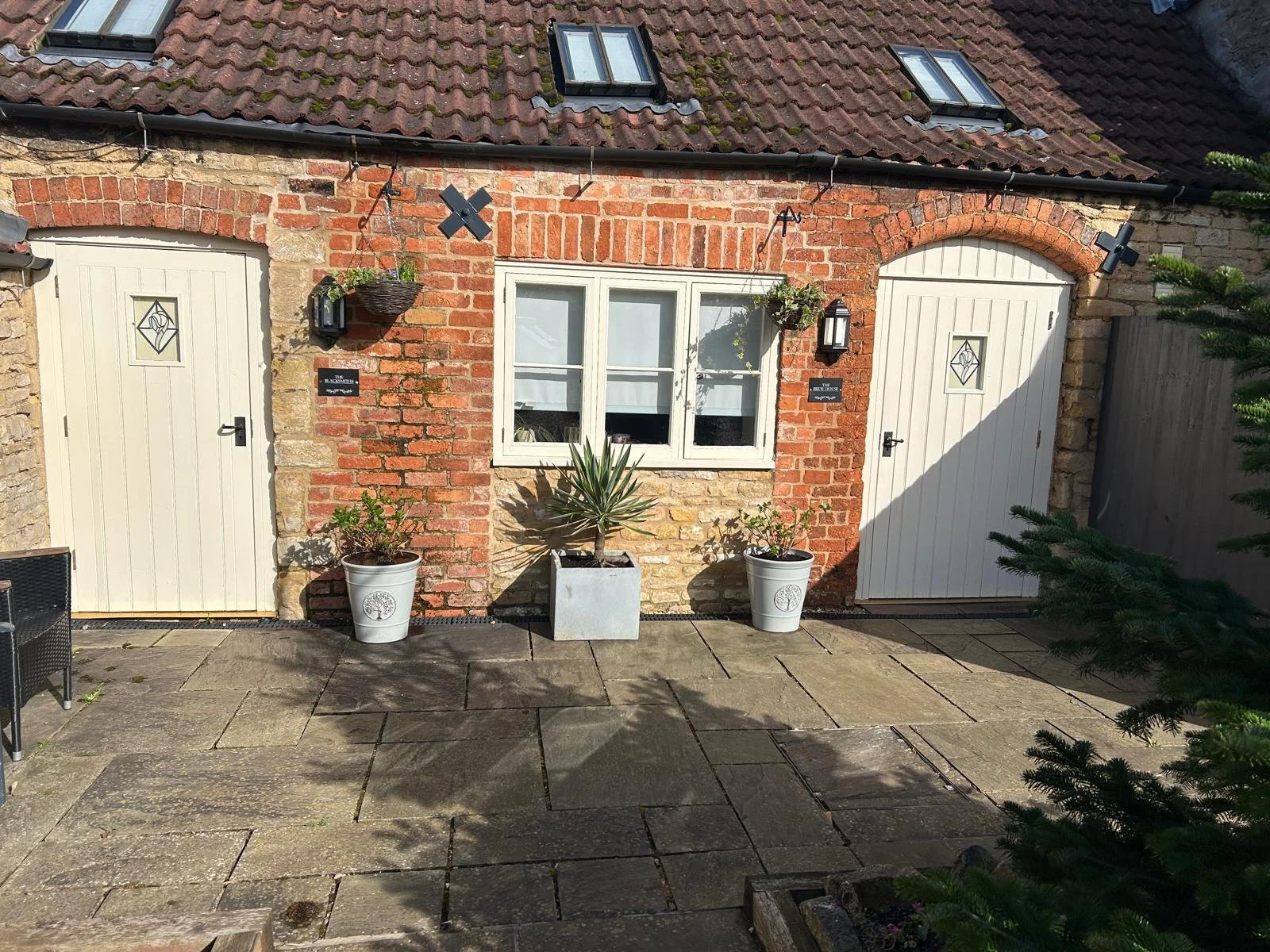 The image shows the exterior of a brick building with a tiled roof and two small windows. The brick wall has two white doors with decorative glass panels, and there are two lantern-style lights on either side. Potted plants are placed in front of the wall, including two white pots with greenery and a central larger planter with a spiky plant. The ground is paved with stone tiles, and part of a tree can be seen on the right side.