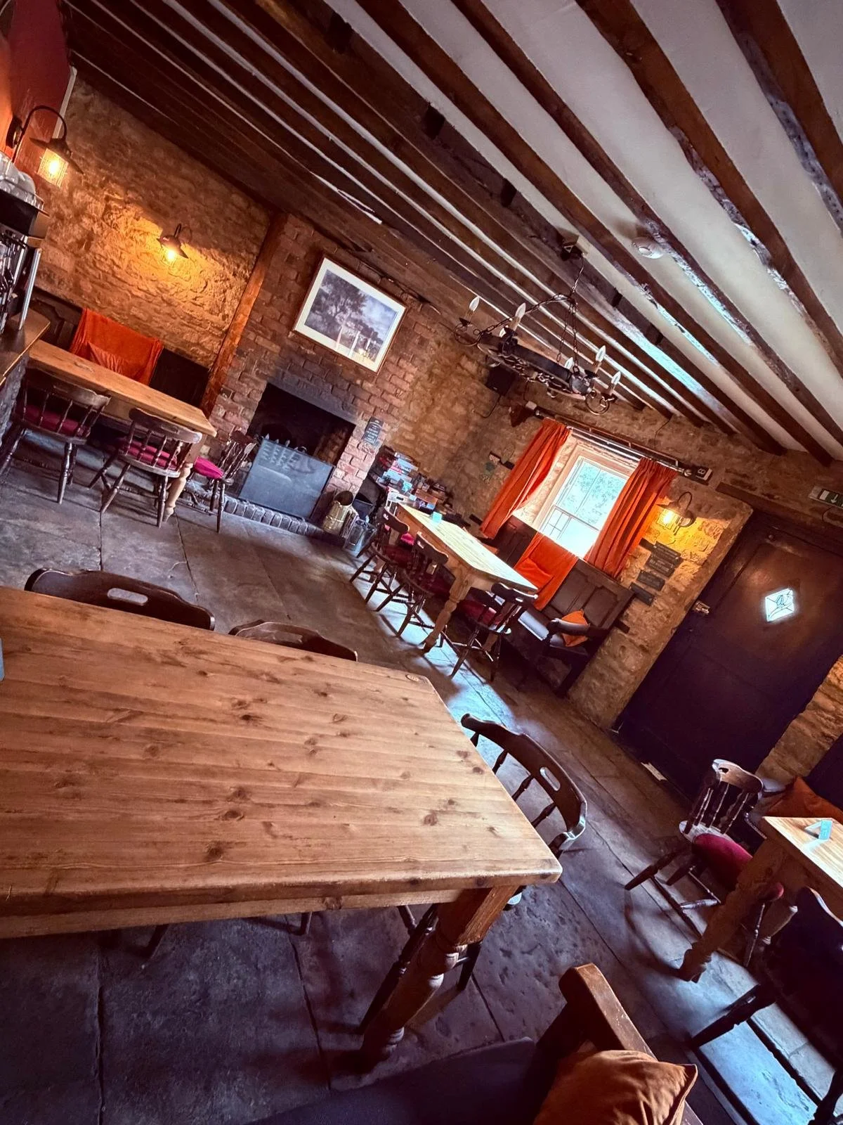 Cozy rustic restaurant interior with exposed brick walls, wooden beams, and warm lighting. Wooden tables and chairs are arranged throughout, with orange curtains and artwork on the walls.