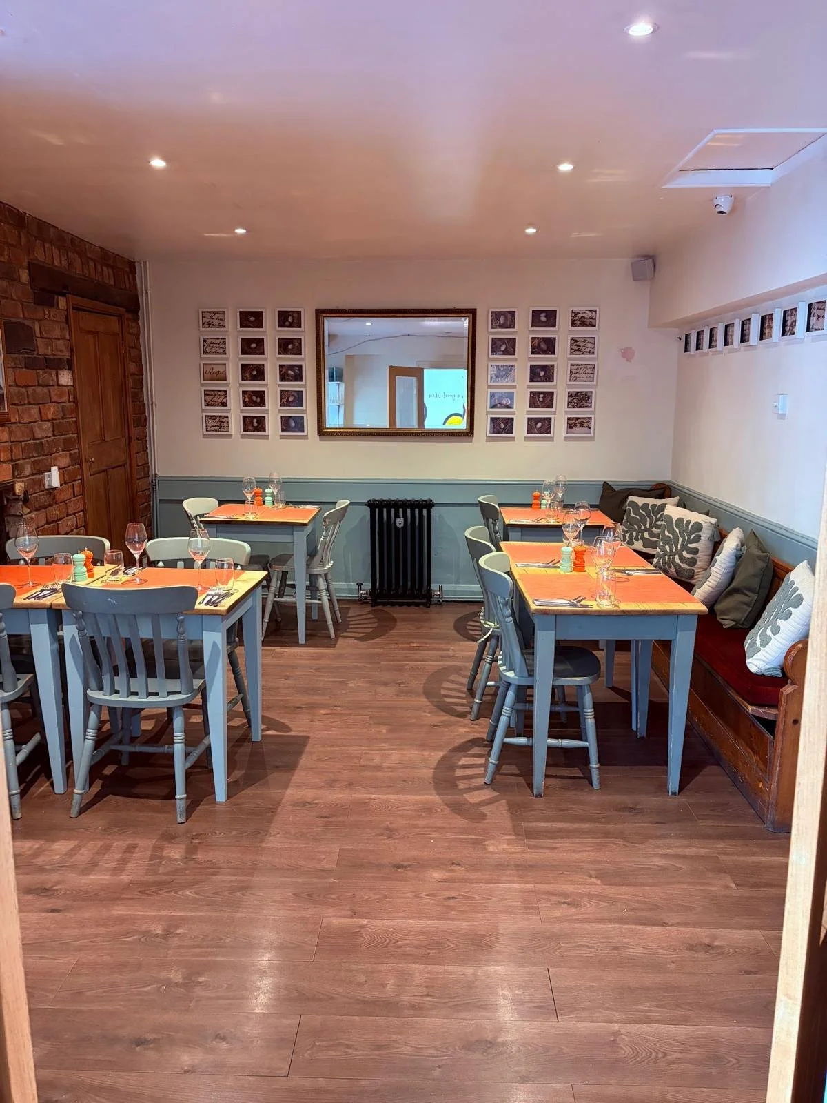 A cozy restaurant with wooden flooring, blue-painted tables, and white chairs. The tables are set with glasses, napkins, and condiments. A study wall decor includes framed pictures and a mirror, with a brick wall on one side and a cushioned seating area along the wall.