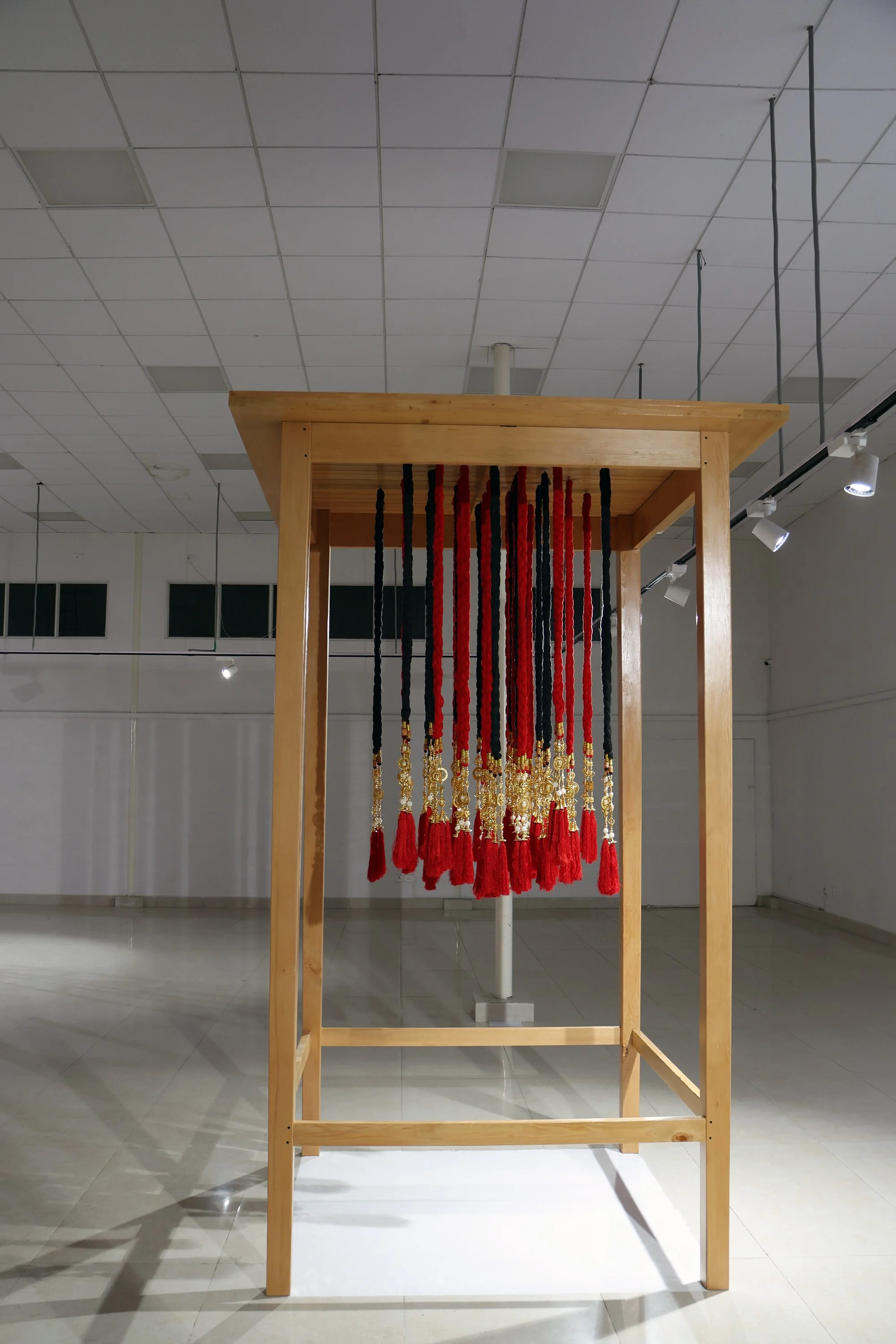 An art installation featuring a wooden frame with hanging red and black tassels, arranged vertically, inside an indoor gallery space with a tiled ceiling and track lighting.