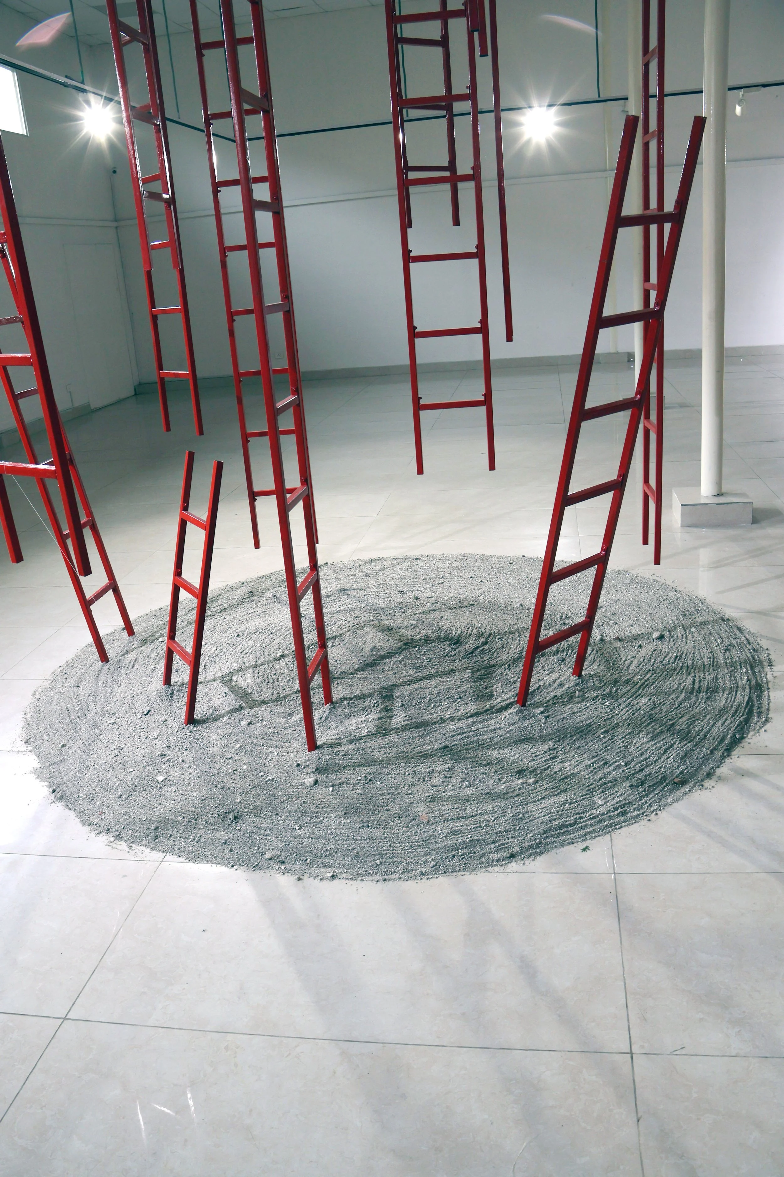 Inside an art gallery or museum, red ladders arranged in a circle on a sand-like circular rug, with white walls, tiled floor, and ceiling lights.