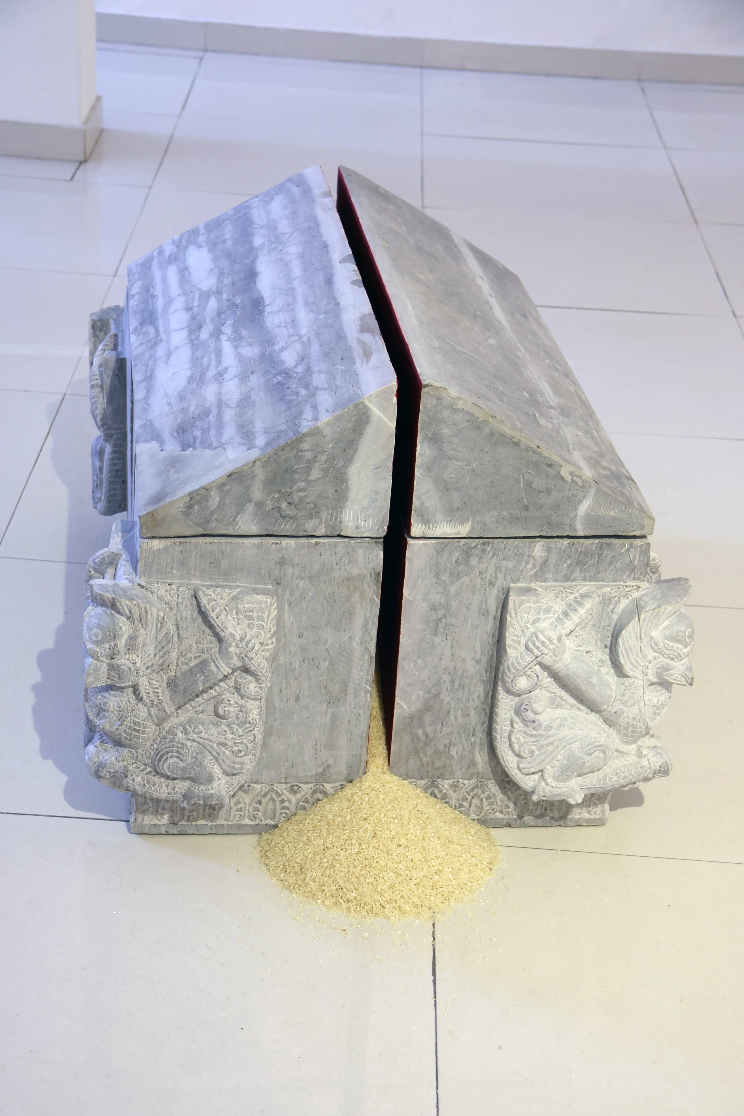 A stone sculpture of two lions with their mouths open, sitting side by side, with a small pile of yellow rice in front of them on a tiled floor.