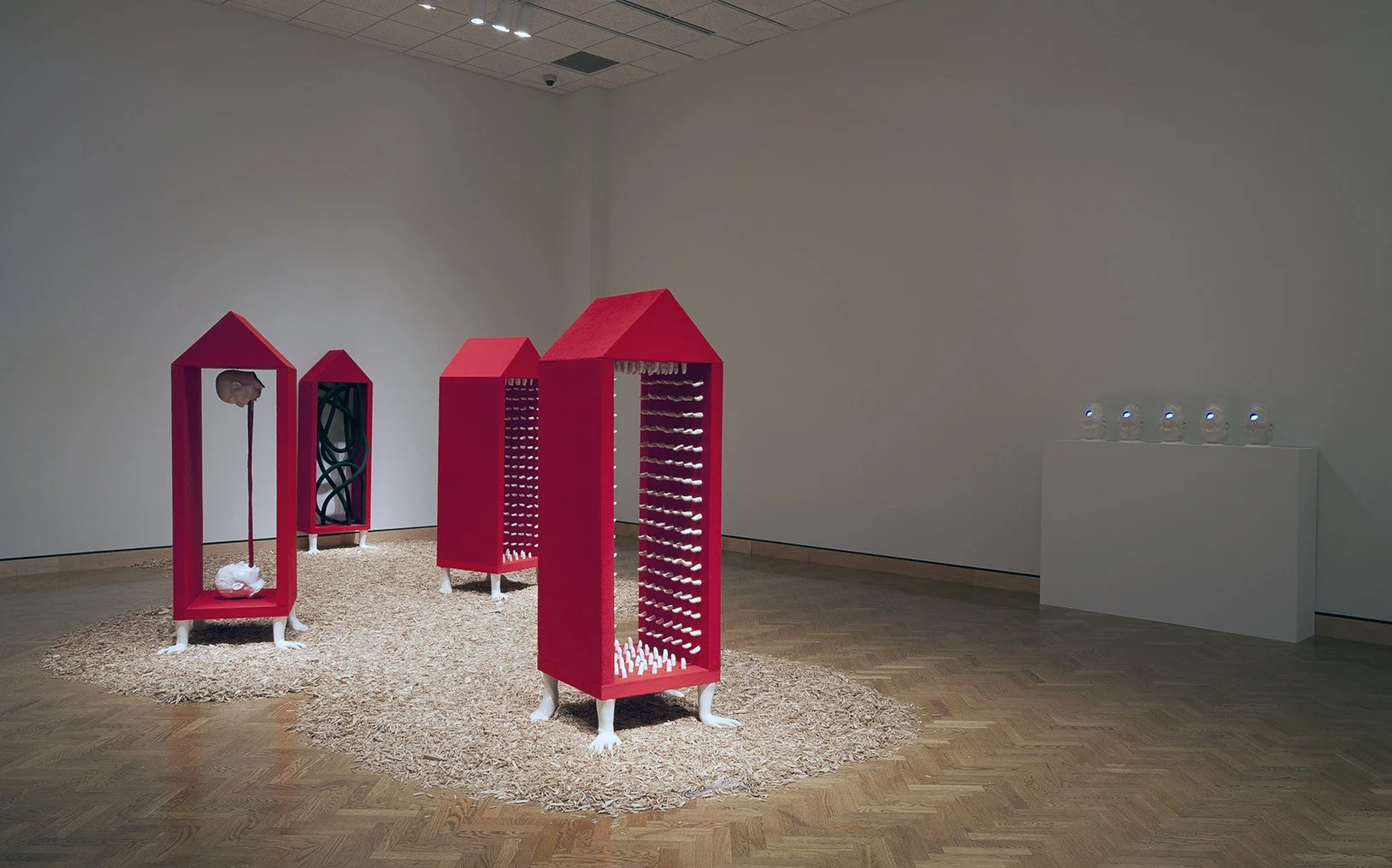 Art installation featuring five vertical structures with red roofs and white legs, resembling small houses, arranged on a circular area covered with beige textured material in an art gallery. Each structure has different internal elements, with some 