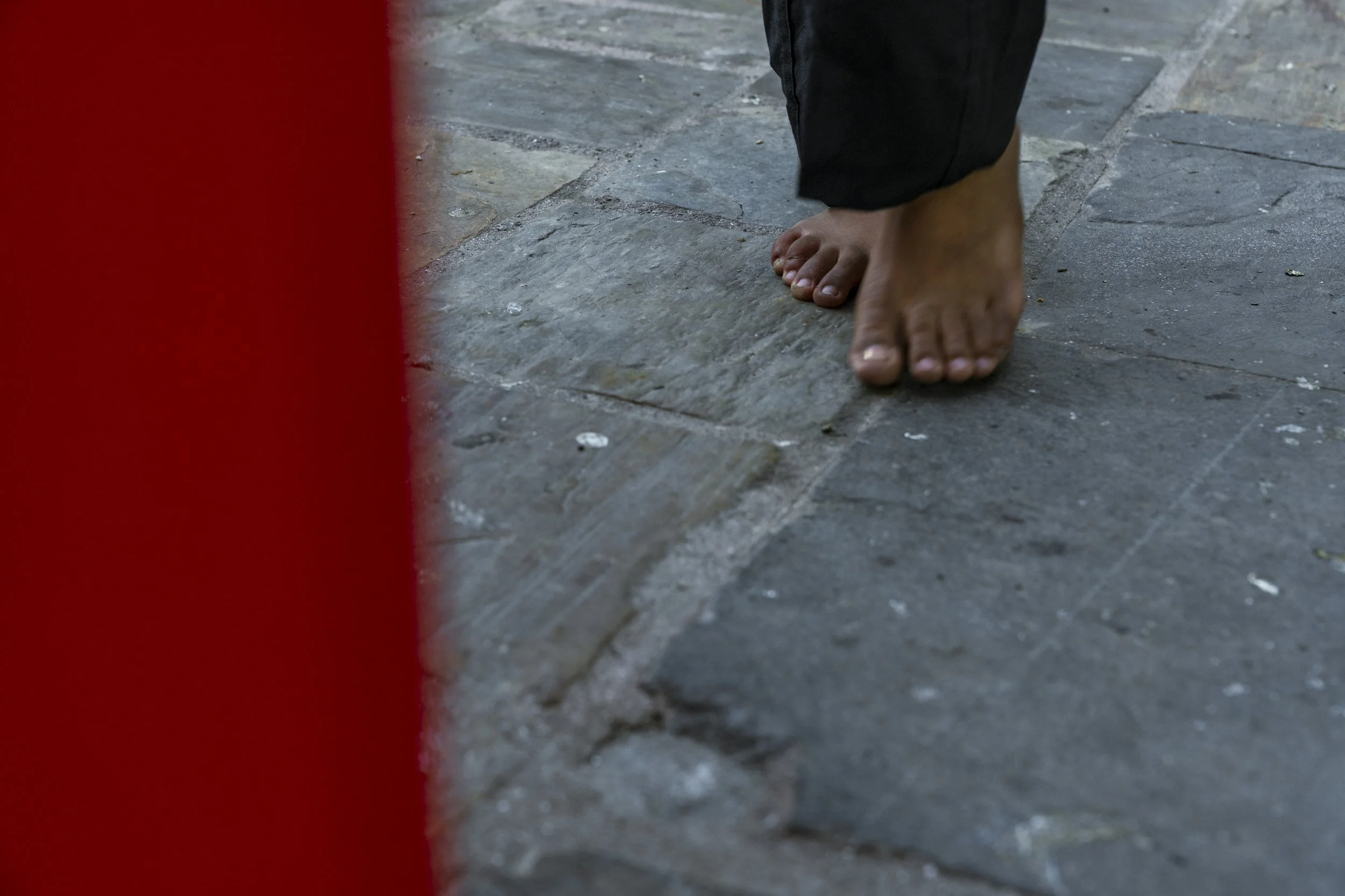 Close-up of a person's bare feet on a stone pavement, with part of a red object or structure on the left side of the image.