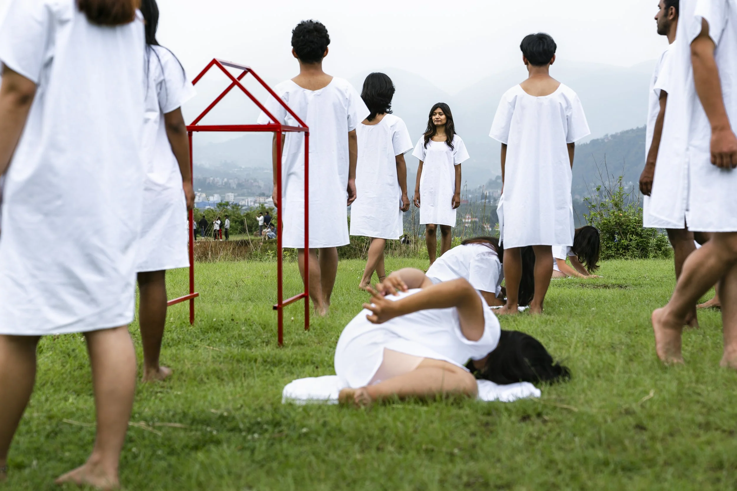 Group of people dressed in white robes standing and sitting on grass outdoors with mountains in the background.