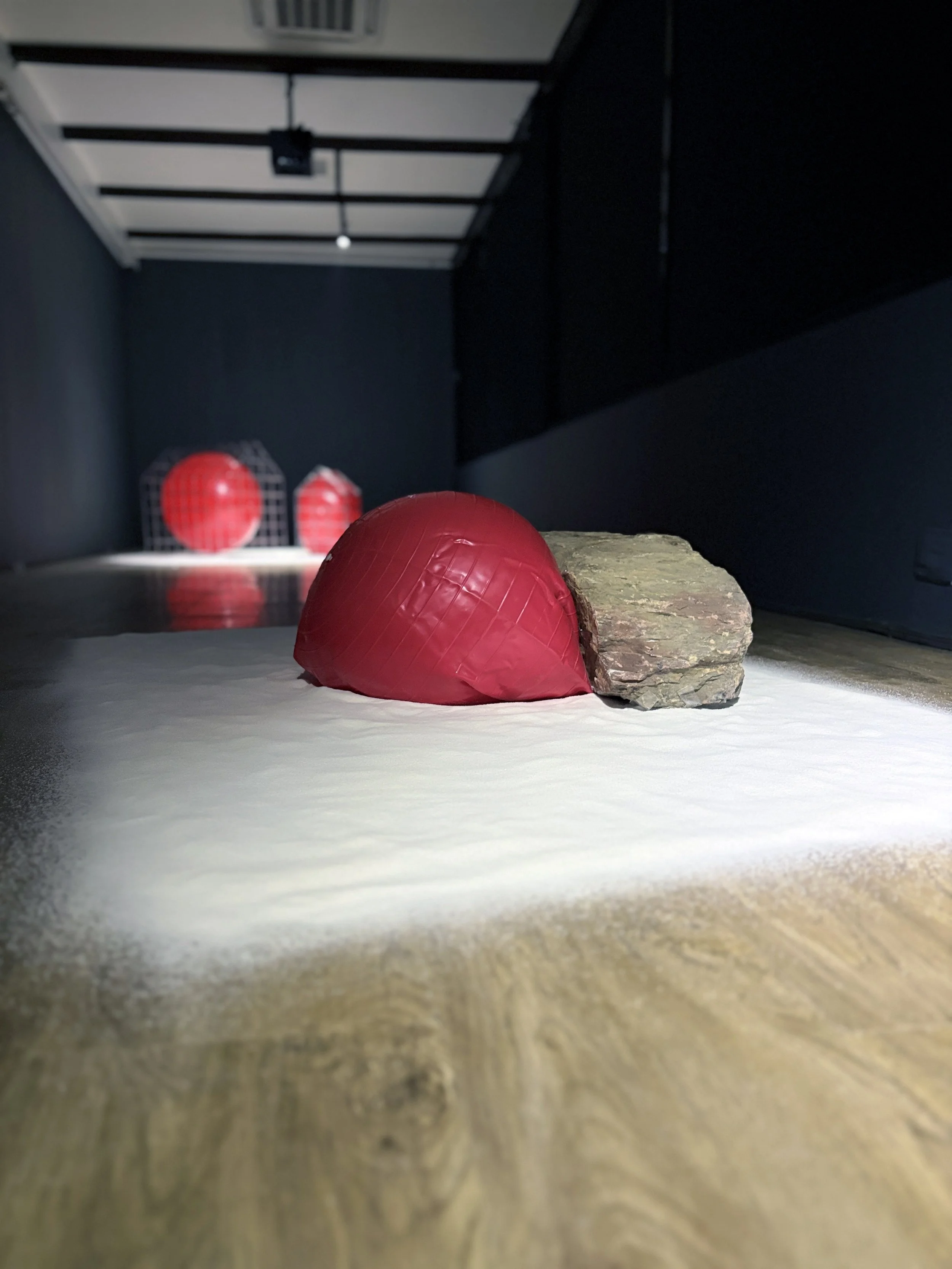 Close-up of a red ball on a light-colored surface with a large rock and two red objects with black grid patterns in the background.