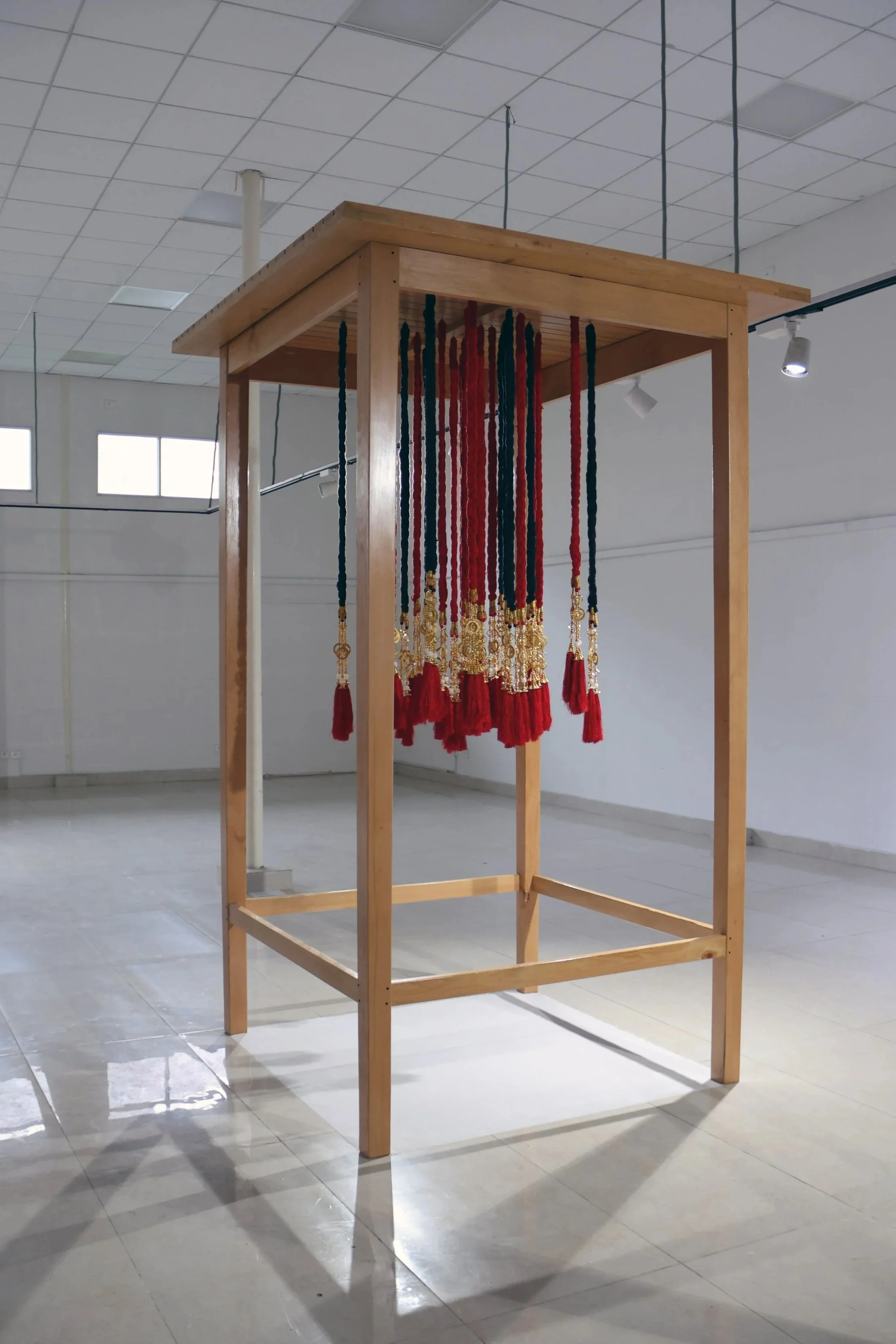A wooden structure displaying red and black braided cords with tassels hanging from the top, set in a large, well-lit room with white walls and ceiling tiles.