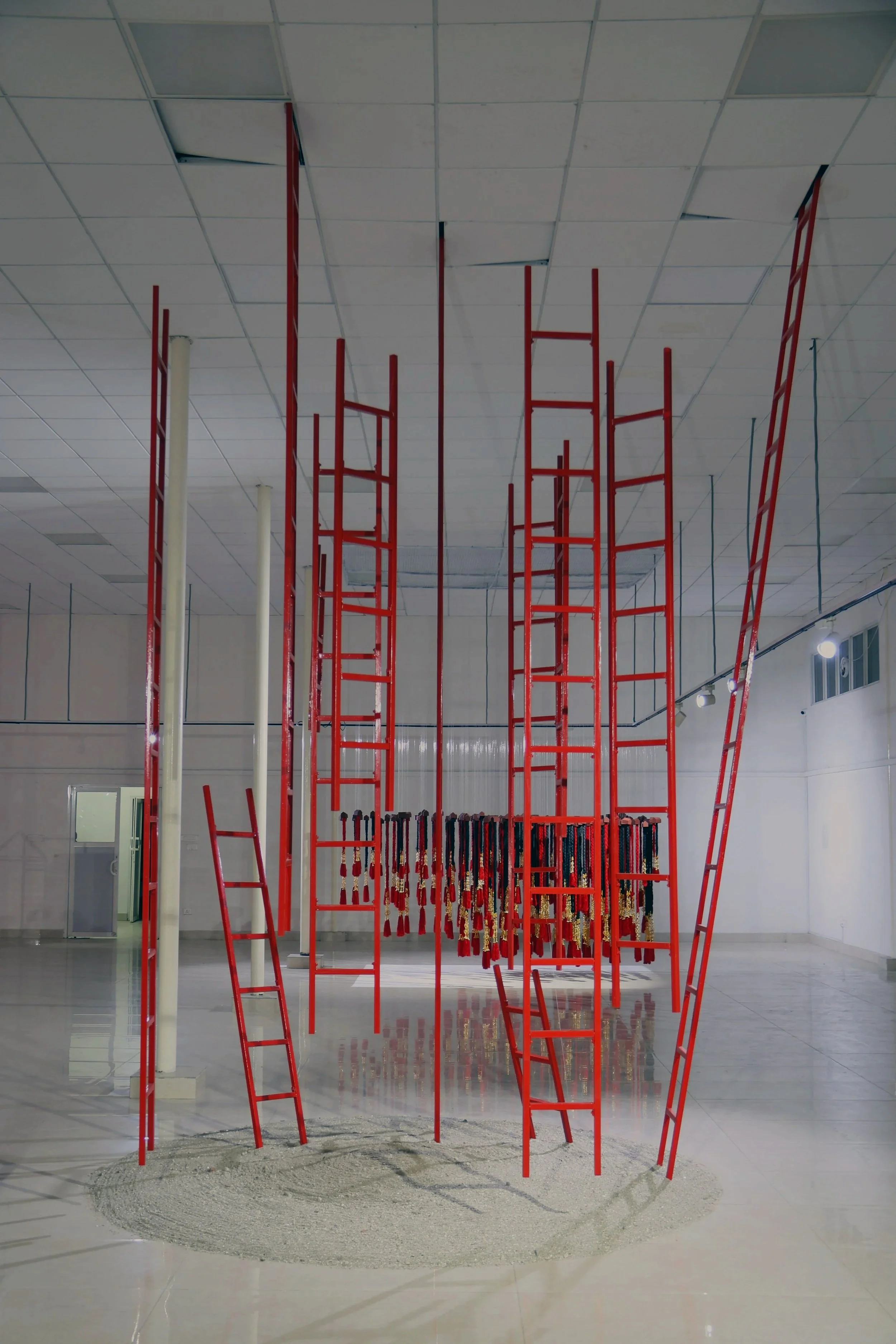 An art installation featuring red ladder-like structures arranged in a circular pattern inside a gallery space. The installation has a base of sand or gravel, reflecting in the polished floor. Hanging from the ceiling are smaller red and black object