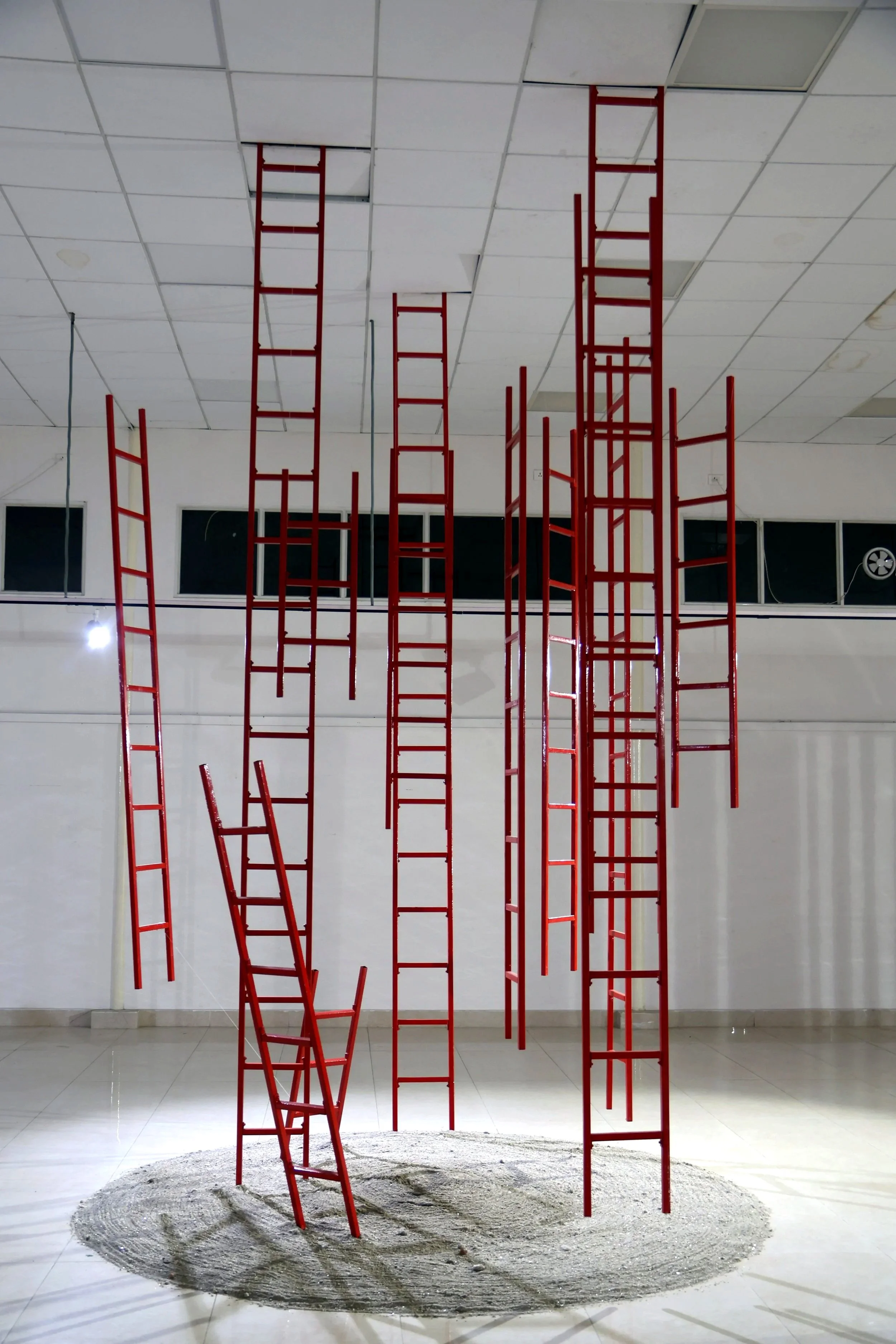 Installation art with red ladders standing on a round patch of sand or dirt in a room with white tiled floors and ceiling. The ladders are tall and various in height, some leaning and others standing upright, creating an abstract composition.