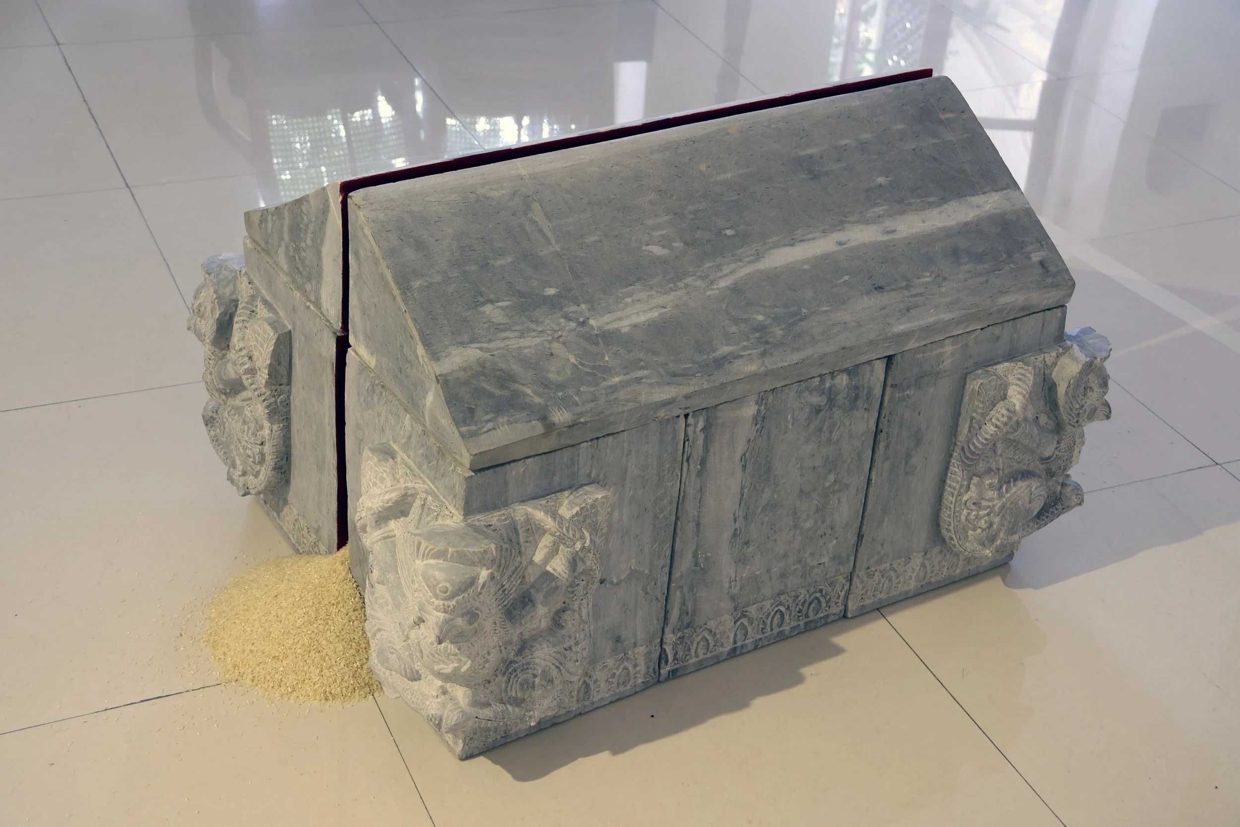 A marble carved structure resembling a small shrine or altar, with detailed decorative carving on the sides and a stepped base, situated on a tiled floor with a pile of sand nearby.