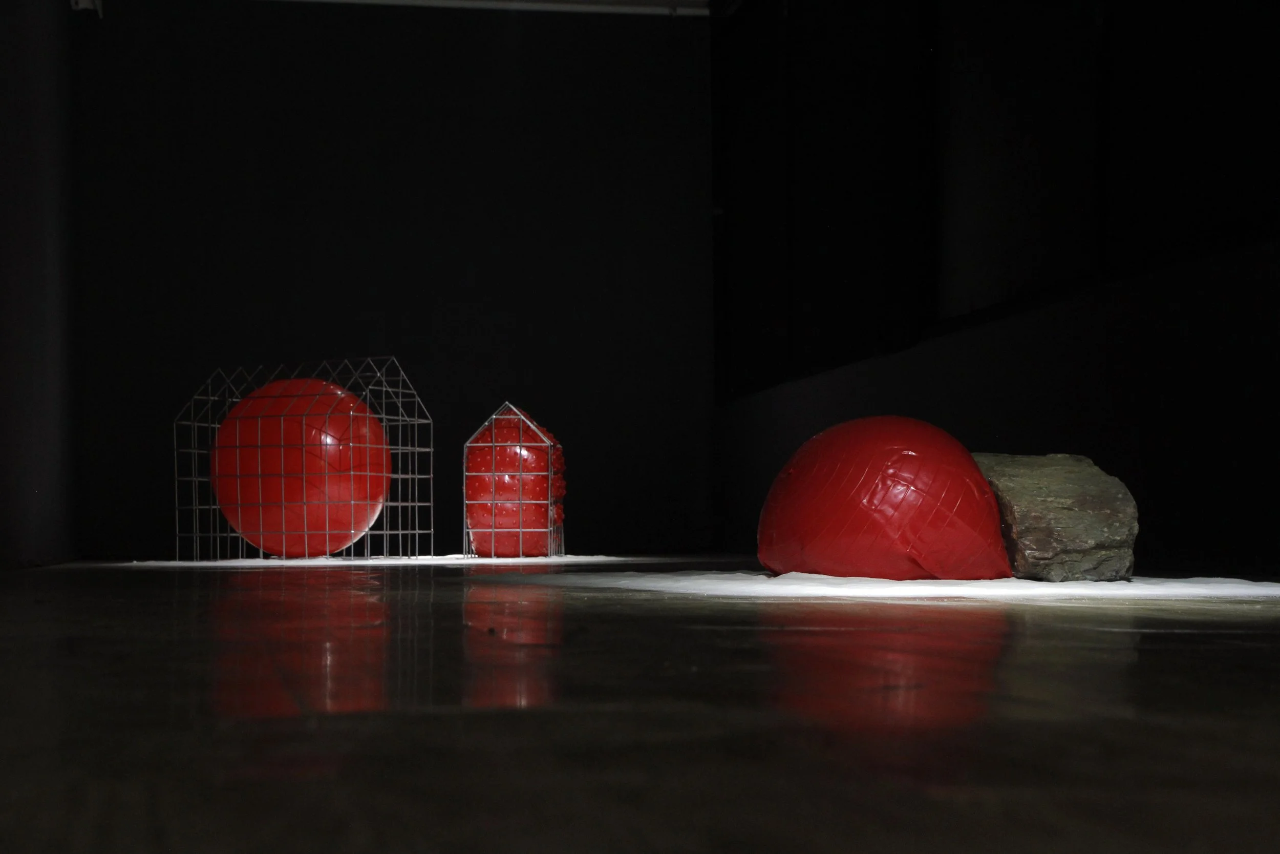 Display of red balls with geometric wire frames and a large rock on the right, on a reflective surface under a spotlight against a dark background.