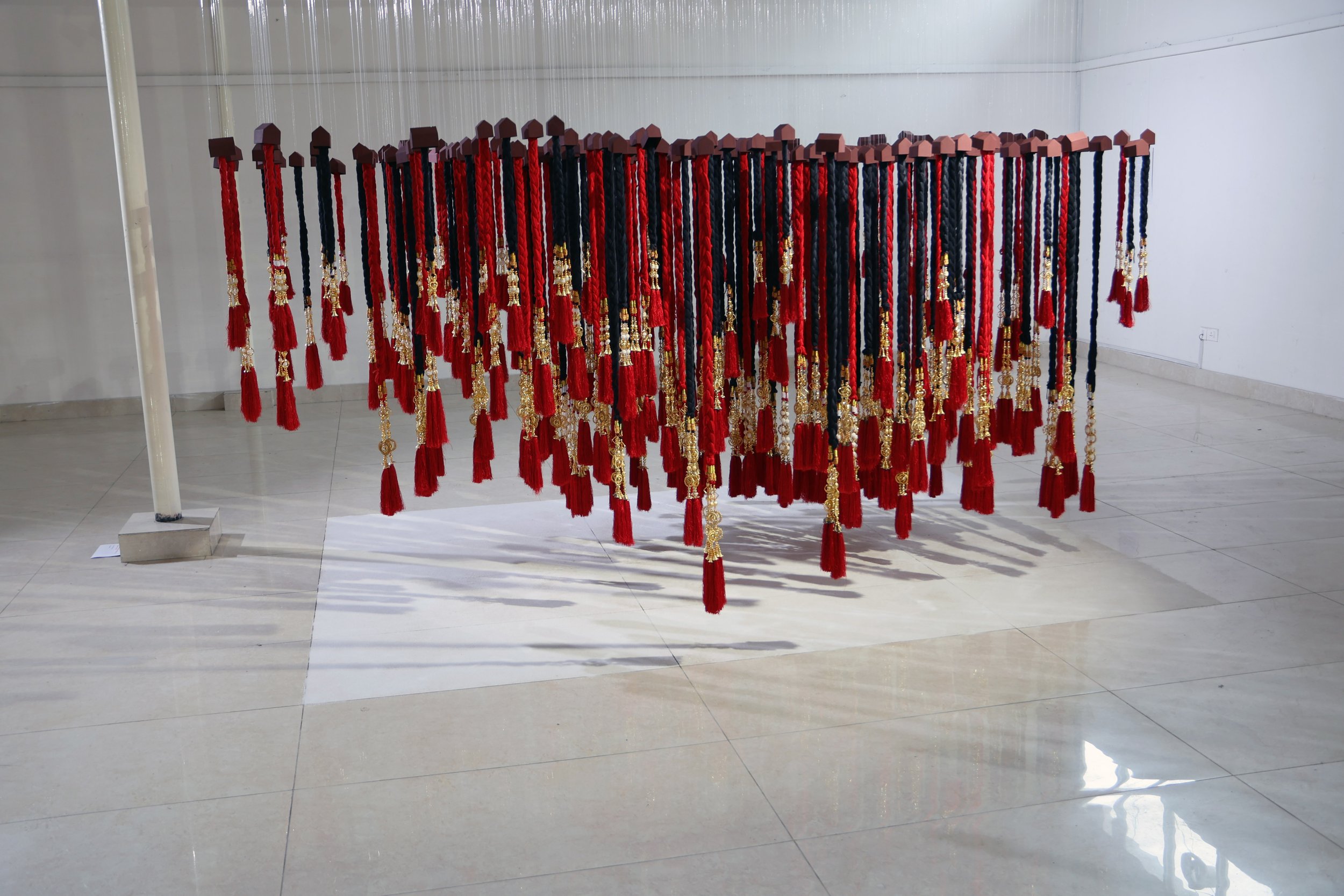 Installation of numerous red and black braided cords hanging from wooden blocks, adorned with gold accents, in a white gallery space.