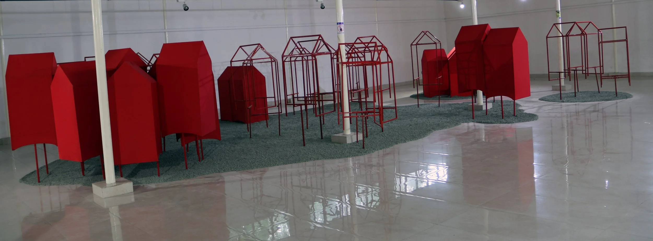 A collection of red abstract art sculptures on a reflective polished floor, arranged on green textured patches, with some sculptures made of metal rods and some of solid red panels in an indoor gallery space.