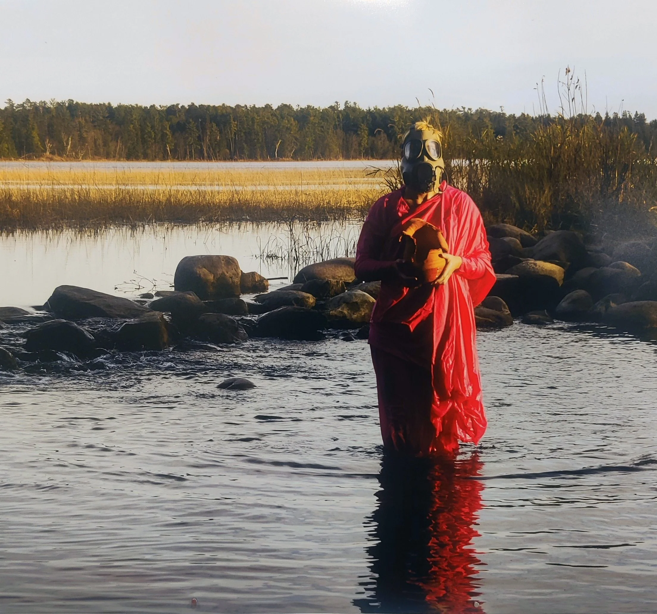 A person wearing a gas mask and a red jacket is sitting on rocks near a body of water, with a line of trees in the background during sunset or sunrise.