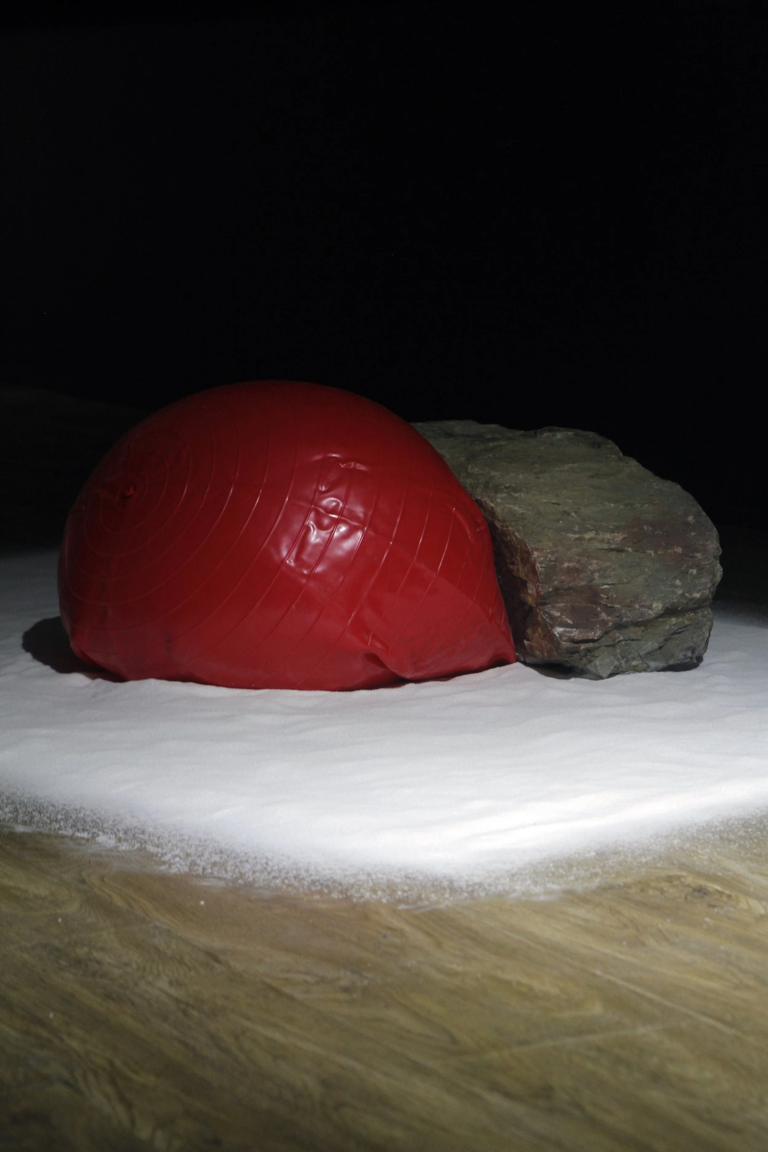 A red exercise ball resting against a large rock on a surface that appears to be snow or salt, with a dark background.