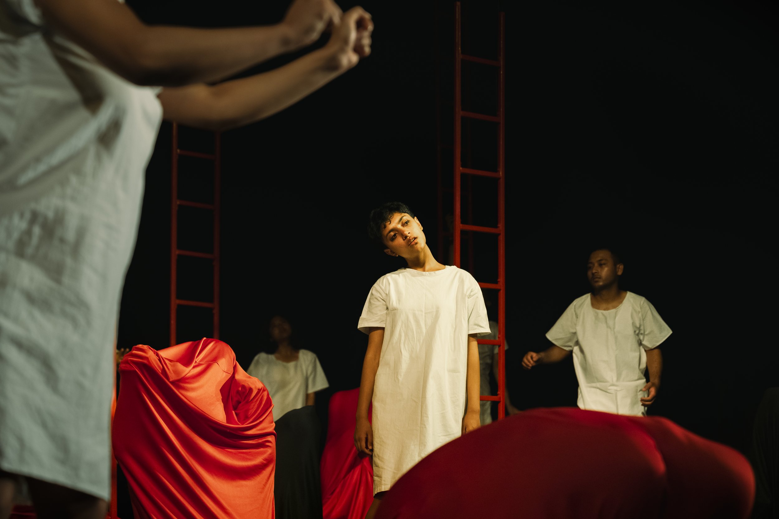 Performers in white dresses stand amidst a group of people, some wearing white shirts and red clothing, in a dark setting with red structural elements visible in the background.