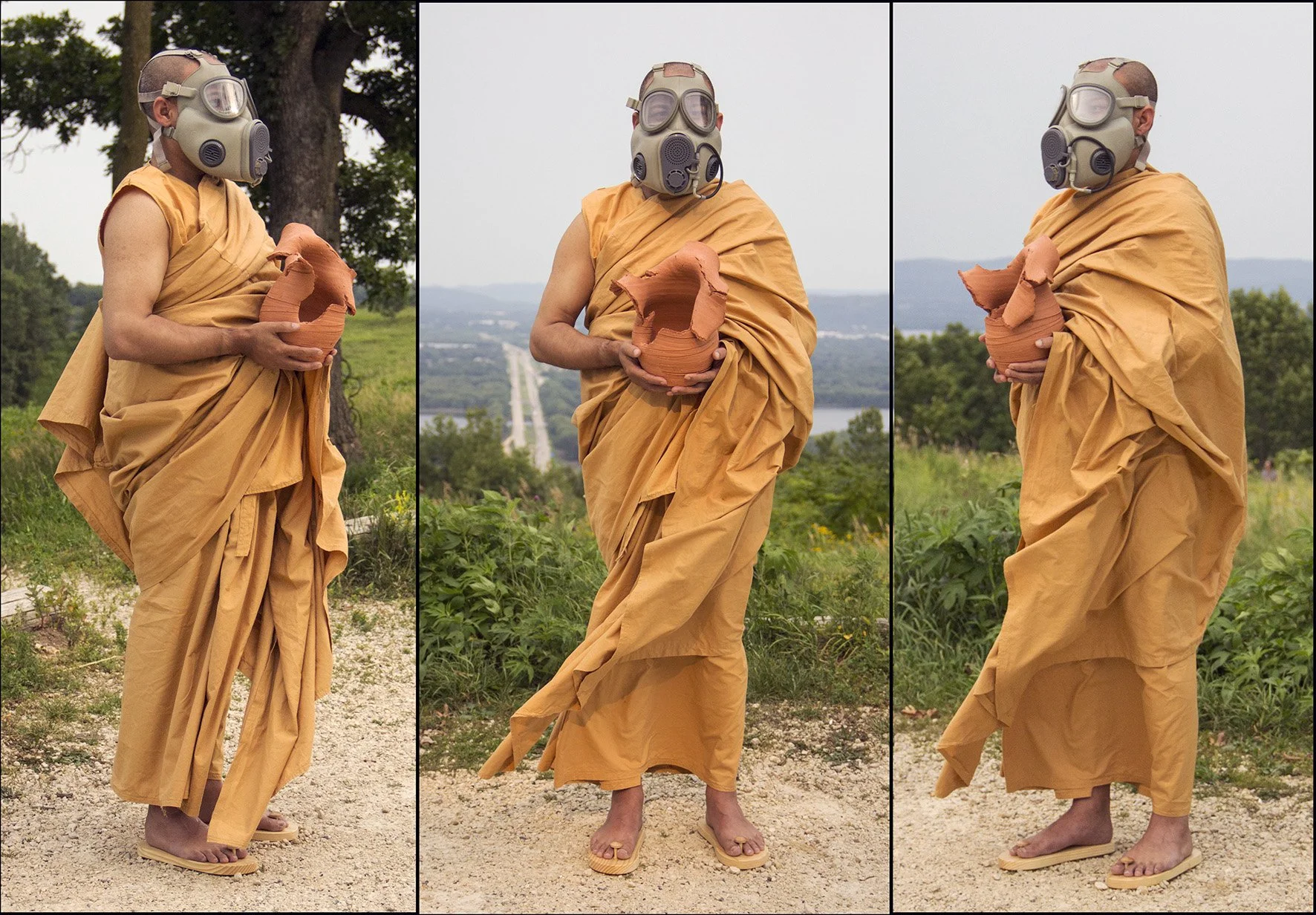 A person dressed in orange Buddhist robes, wearing a gas mask with large eyepieces and filters, standing outdoors on a gravel path. They are holding a broken clay pot in their hands and wearing traditional wooden sandals. The background includes tree
