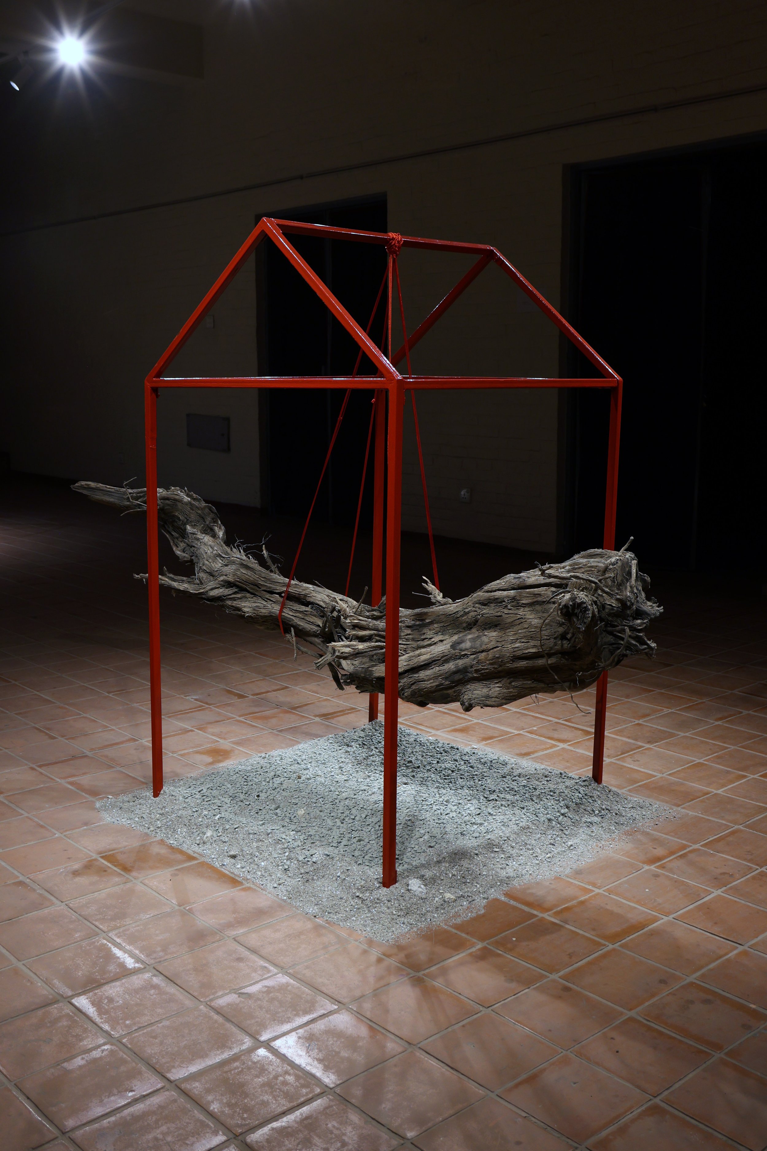 An art installation featuring a large, weathered, twisted piece of driftwood on top of a bed of small gravel. The driftwood is held up by a red metal frame forming a geometric shape. The setting appears to be indoors under dim lighting with a brick w