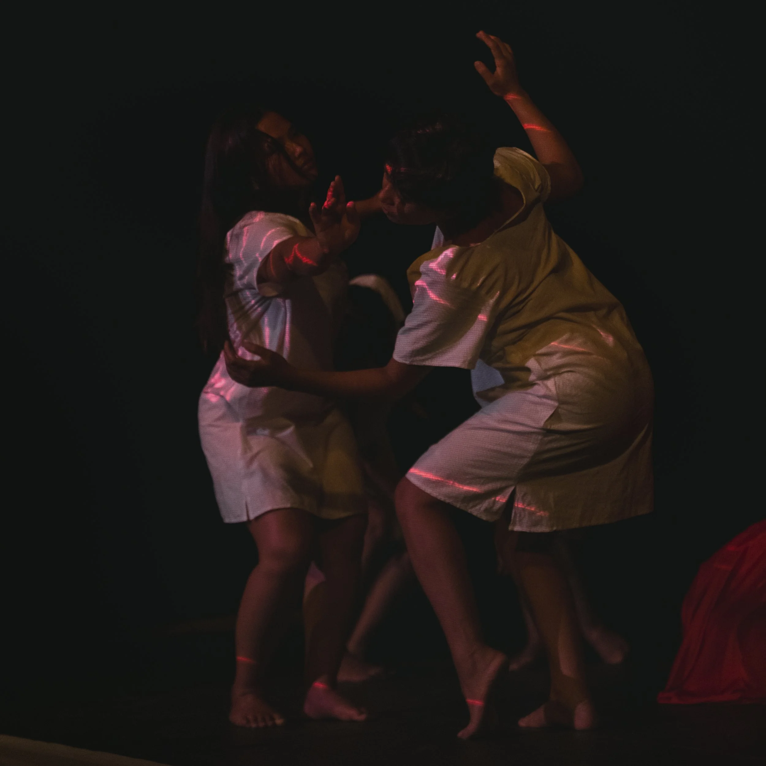 Two performers stand on a dark stage, lit by faint red light.