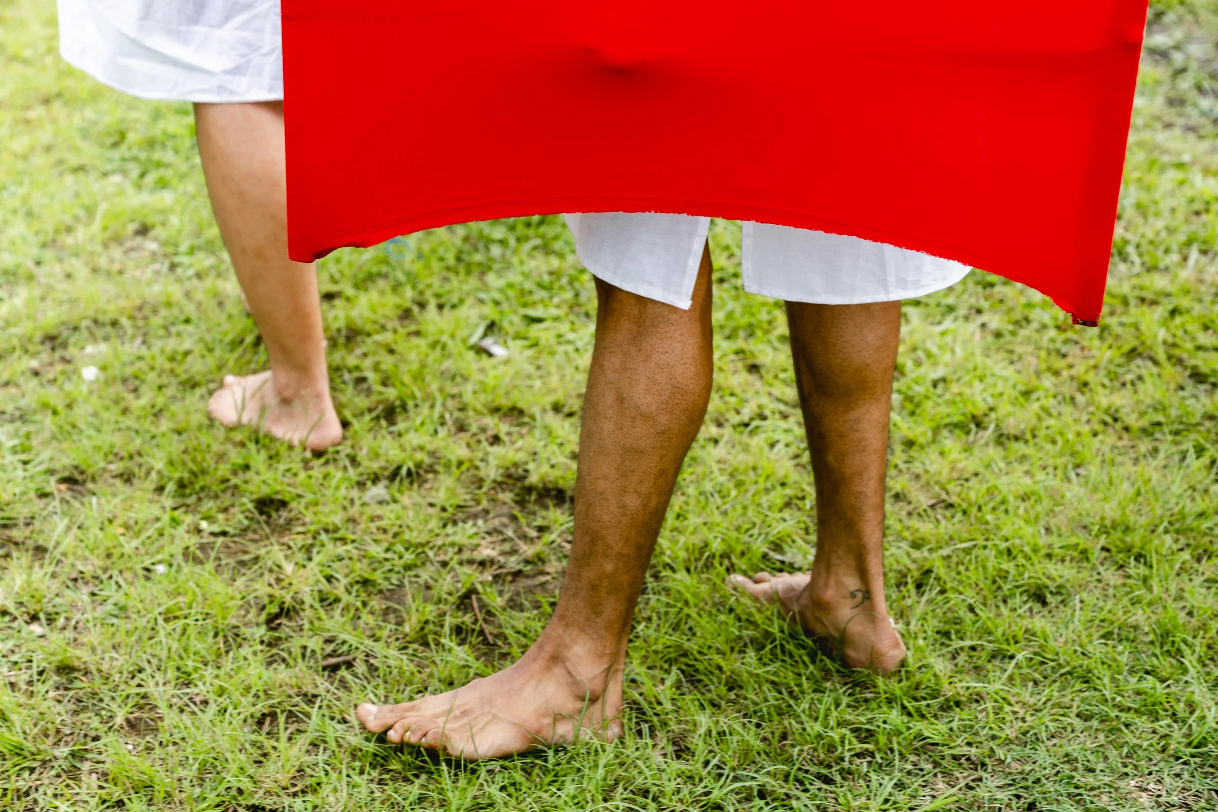 Close-up of two people standing on grass, one barefoot, with visible legs and white clothing, and a person in a white outfit and a red cloth or flag draped over their lower body.