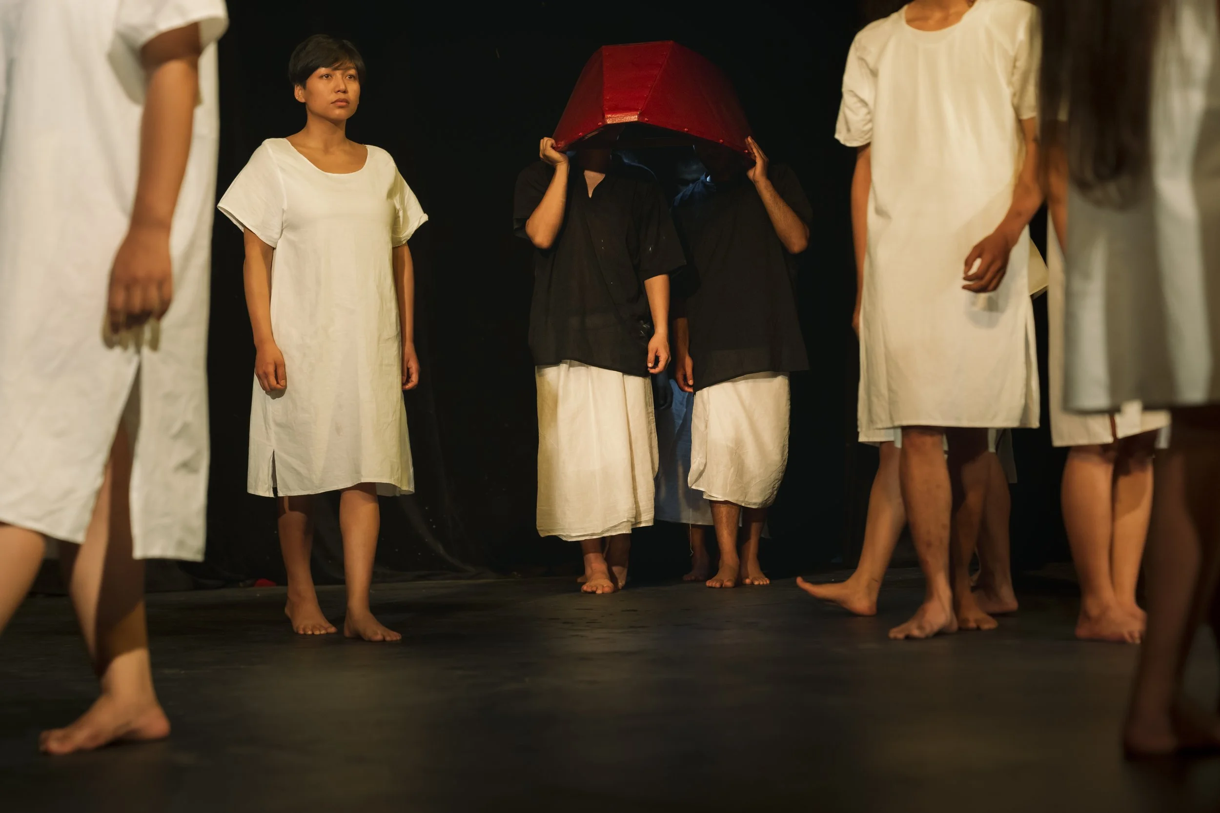 People on stage in traditional costumes, some barefoot, with one person holding a large red head covering, participating in a cultural or theatrical performance.