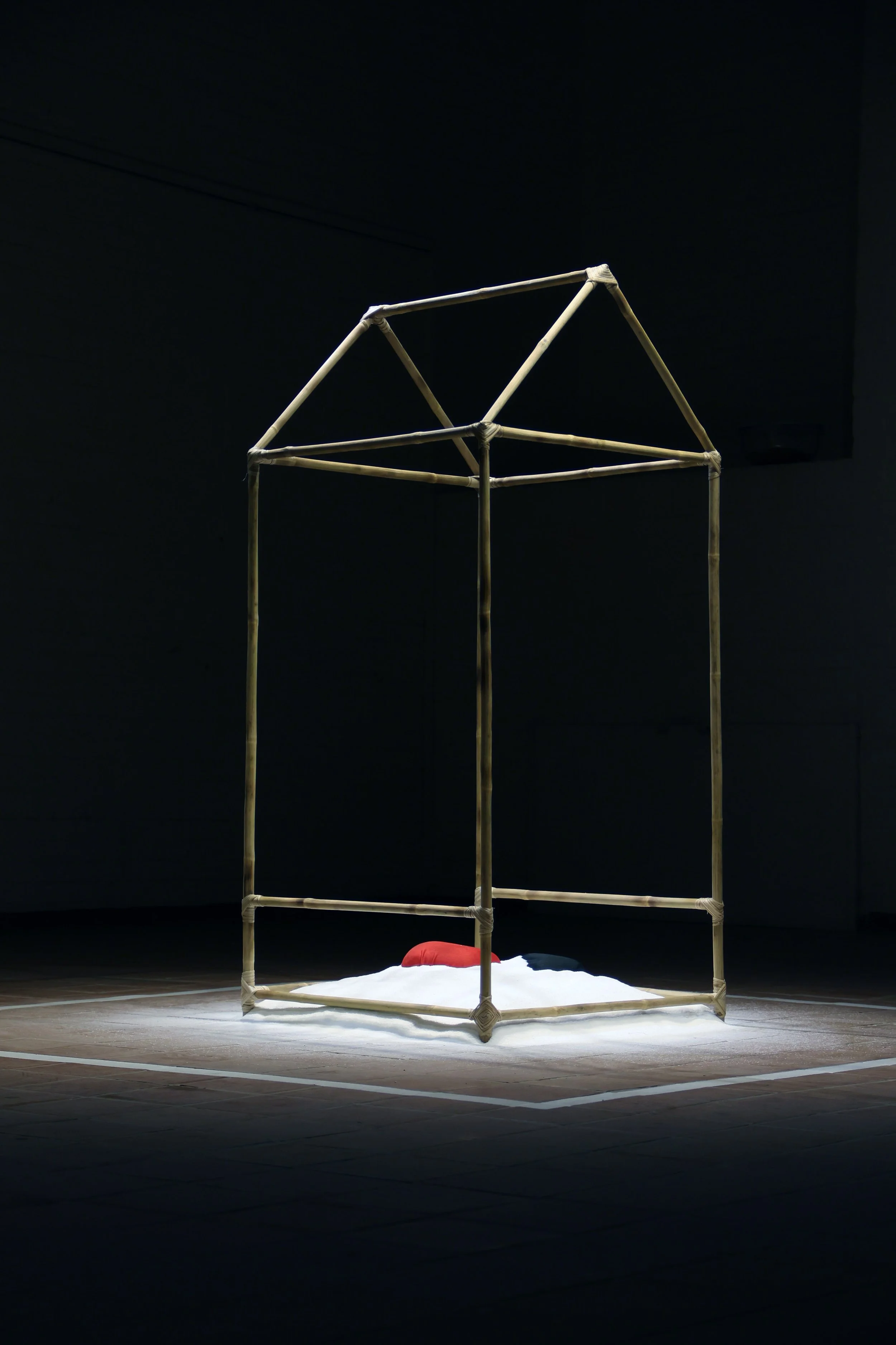 A minimalist art installation features a wooden house-like frame on a stage with a black background, a white cloth underneath, and a red object.