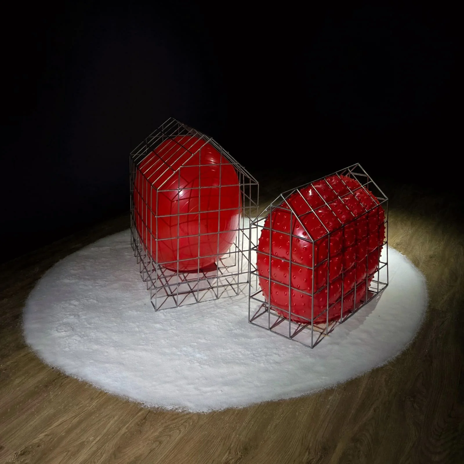 Two art installations of large red objects inside wireframe cages, one shaped like an egg and the other like a rectangular block, displayed on a white surface in a dark gallery setting.