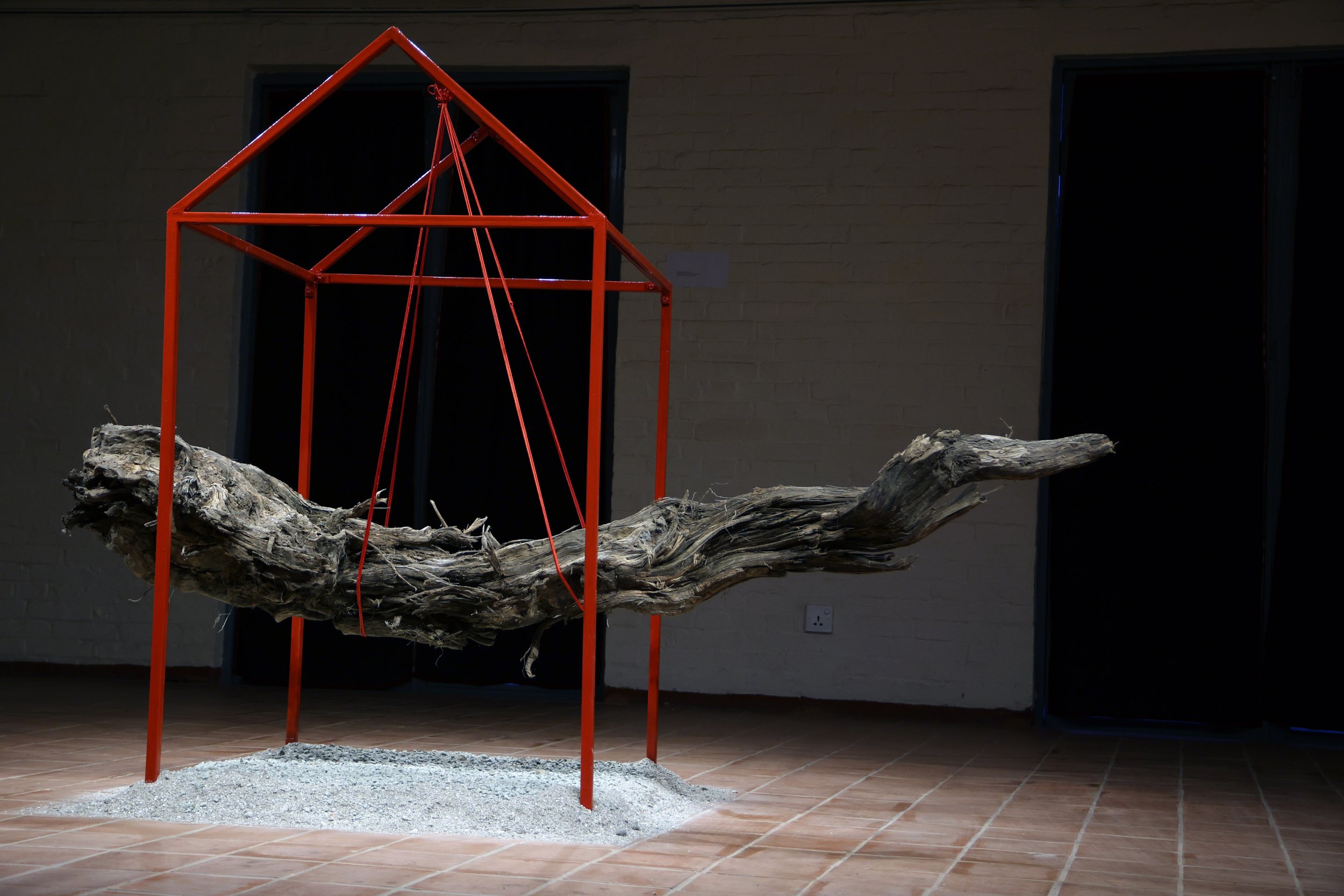 An art installation featuring a large, weathered tree branch supported by a red metal frame with wires, set on a tiled floor with a small pile of gravel underneath.