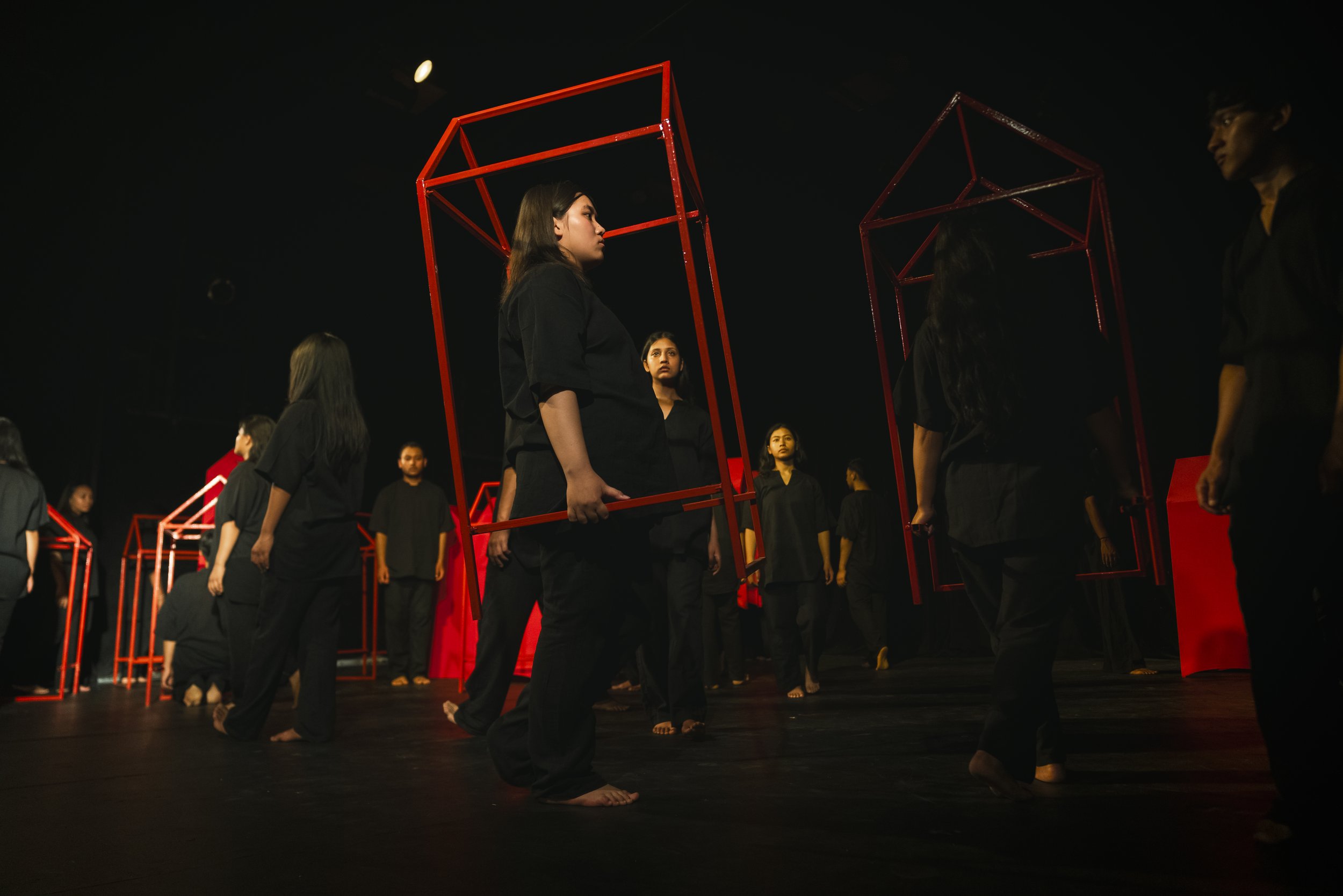 Group of women dressed in black participating in a theatrical performance or rehearsal, holding red metal frames, standing on a dark stage.