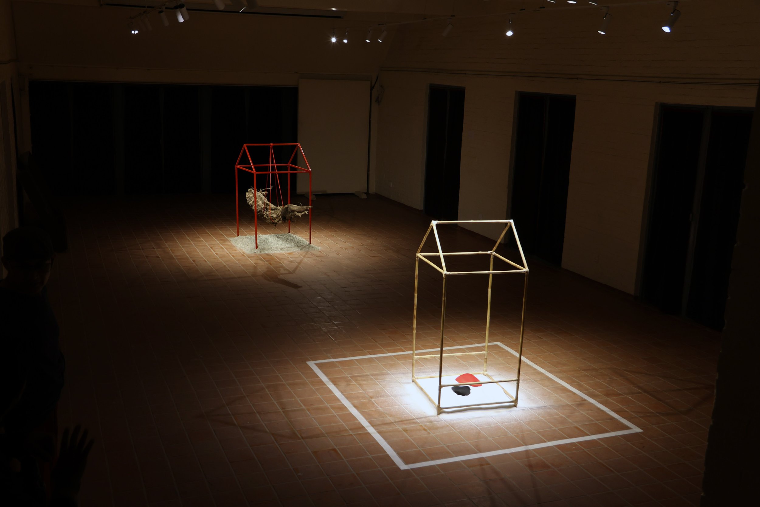 An art installation with geometric metal structures on a tiled floor in a dimly lit room. The structures include a red house-like shape with a hanging fabric and a white 3D cube frame, with a white square marked on the floor around the frame.