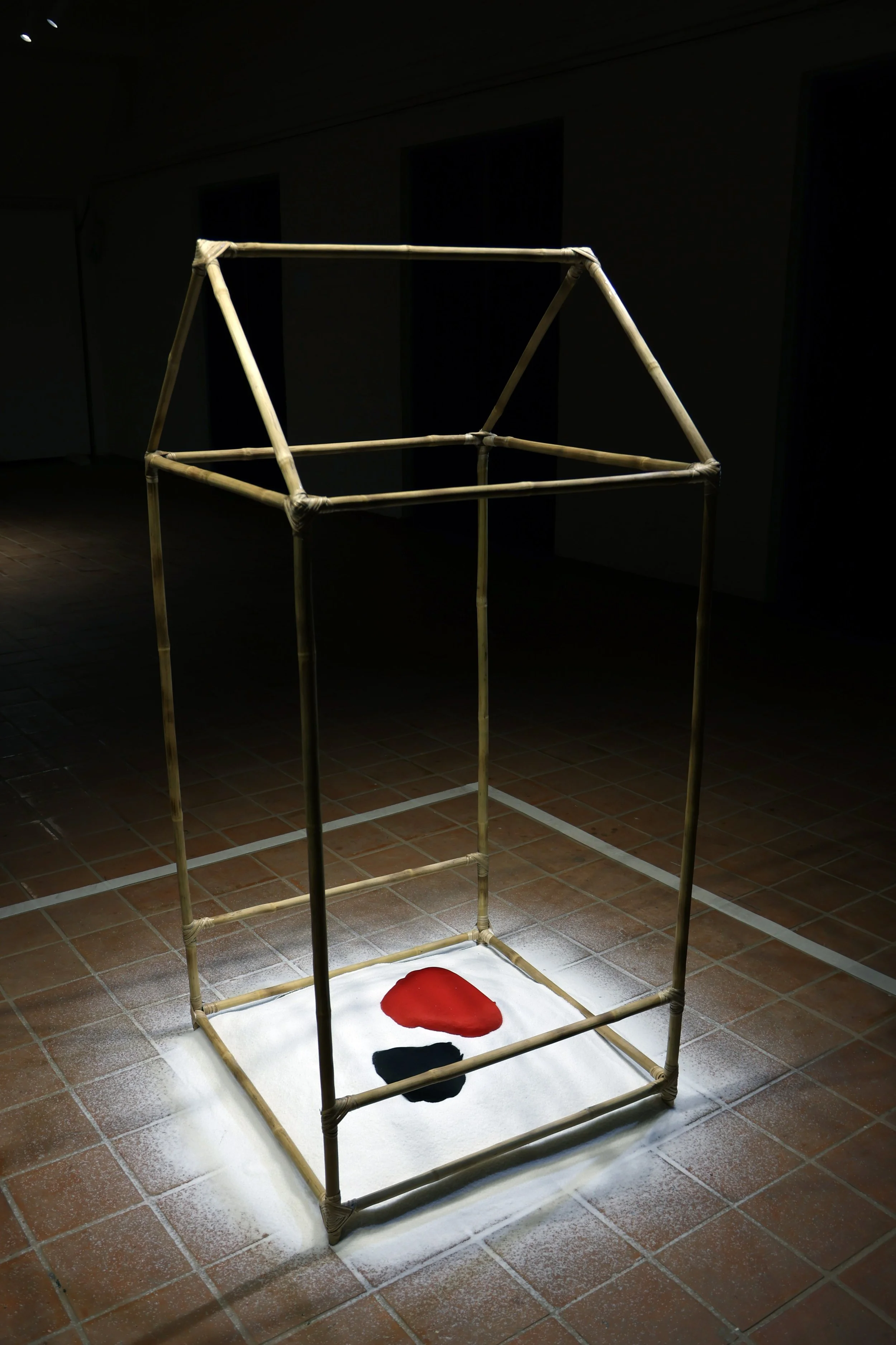An art installation consisting of a wooden frame with a white cloth base on the floor. On the base, there are irregularly shaped red and black objects, arranged in a specific formation. The scene is dimly lit, with a focused light illuminating the ar