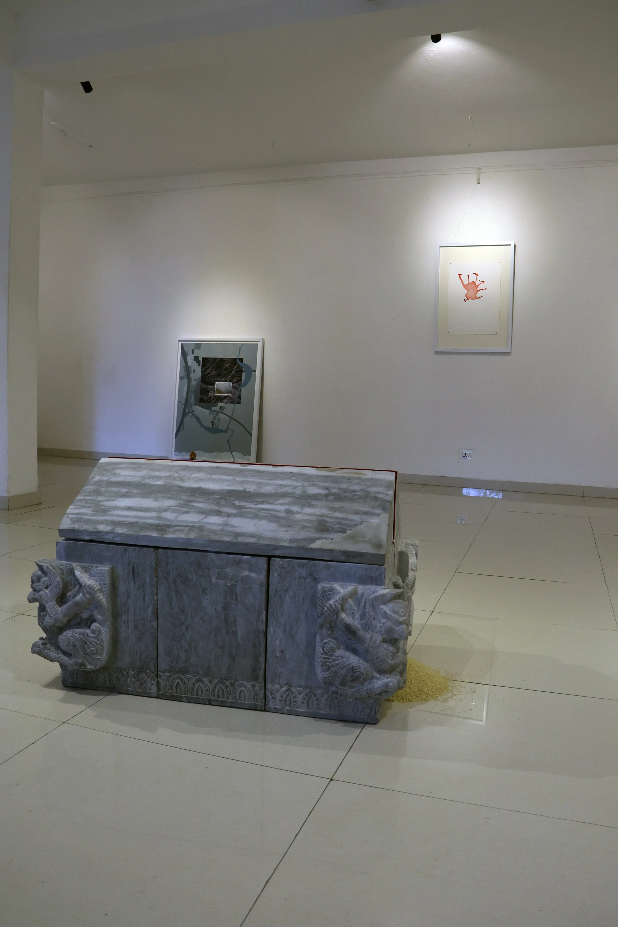A white marble table with detailed carved animal figures on the sides, situated in an art gallery. There are framed artworks on the wall behind, and the gallery has a glossy tiled floor with overhead lighting.