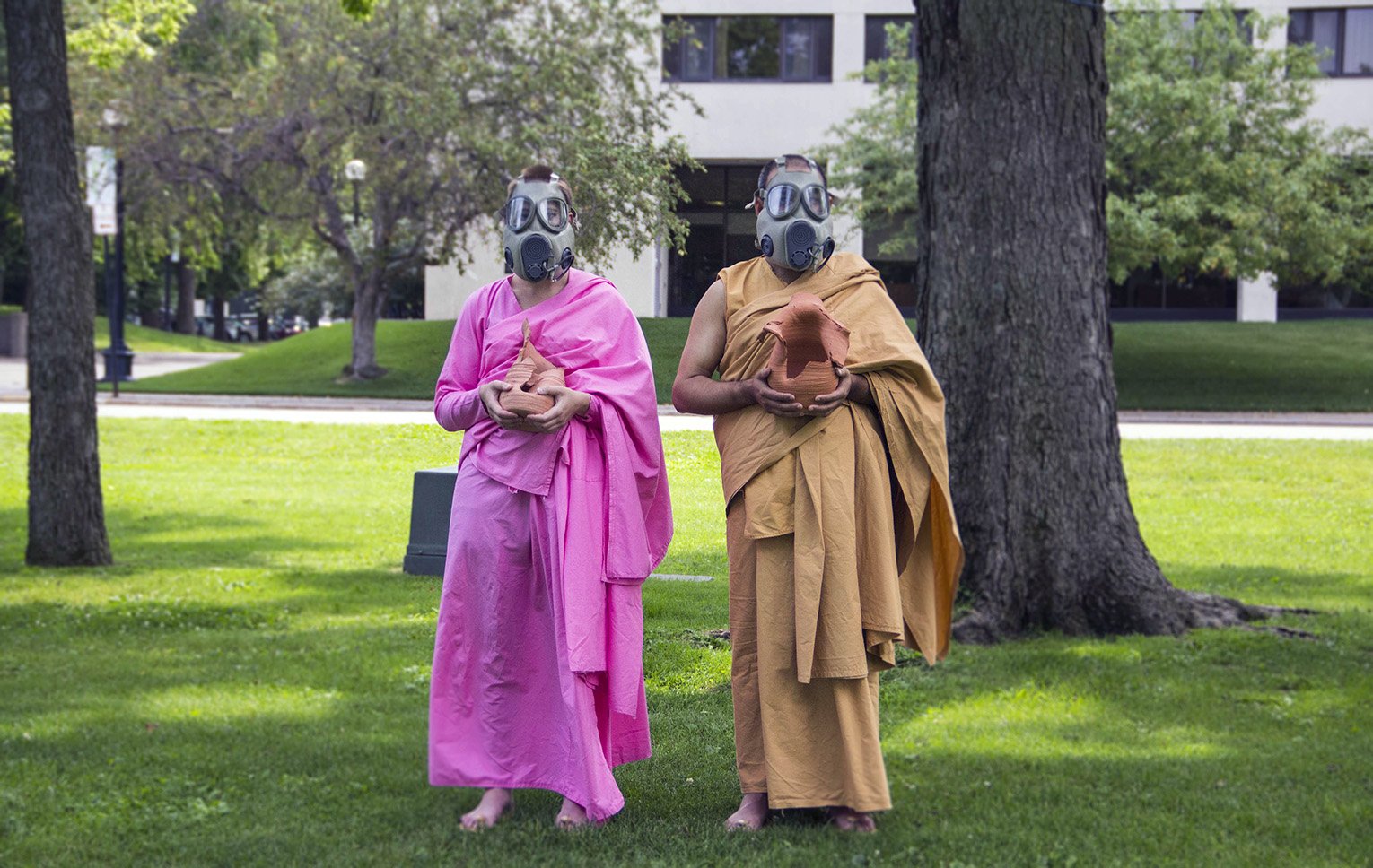 Two people dressed as Buddhist monks wearing masks and robes, holding small clay pots, standing on grass in a park with trees and a building in the background.