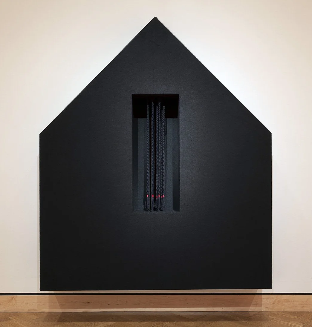 A sculptural work resembling a house with a vertical rectangular cutout in the center, displaying hanging black ropes with red accents inside.