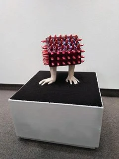 Sculpture of a figure with a body made of animal legs and a torso covered with red and purple spikes, displayed on a black pedestal.