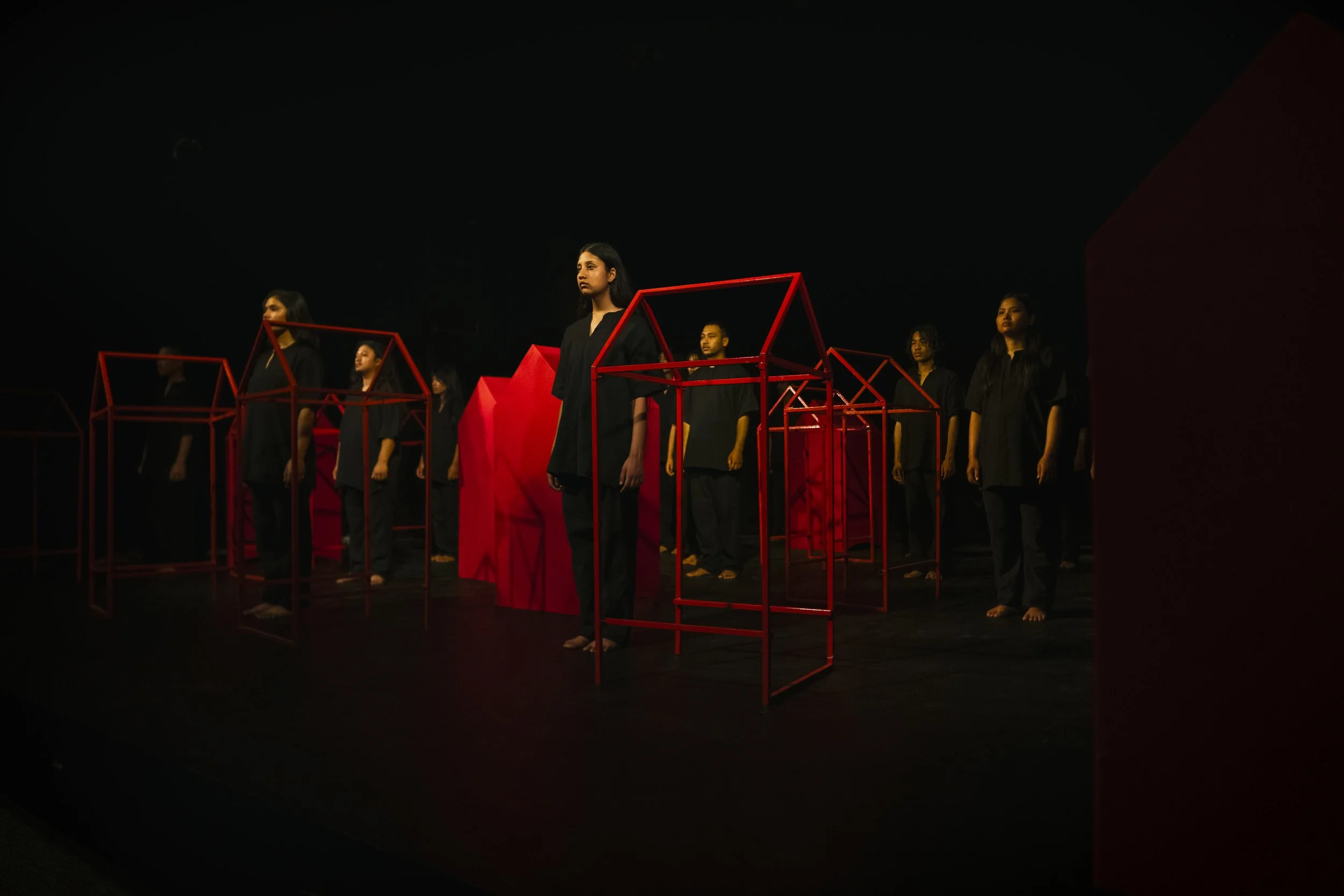 Performers stand in a line on a dark stage, wearing black outfits and serious expressions, surrounded by red geometric frames.