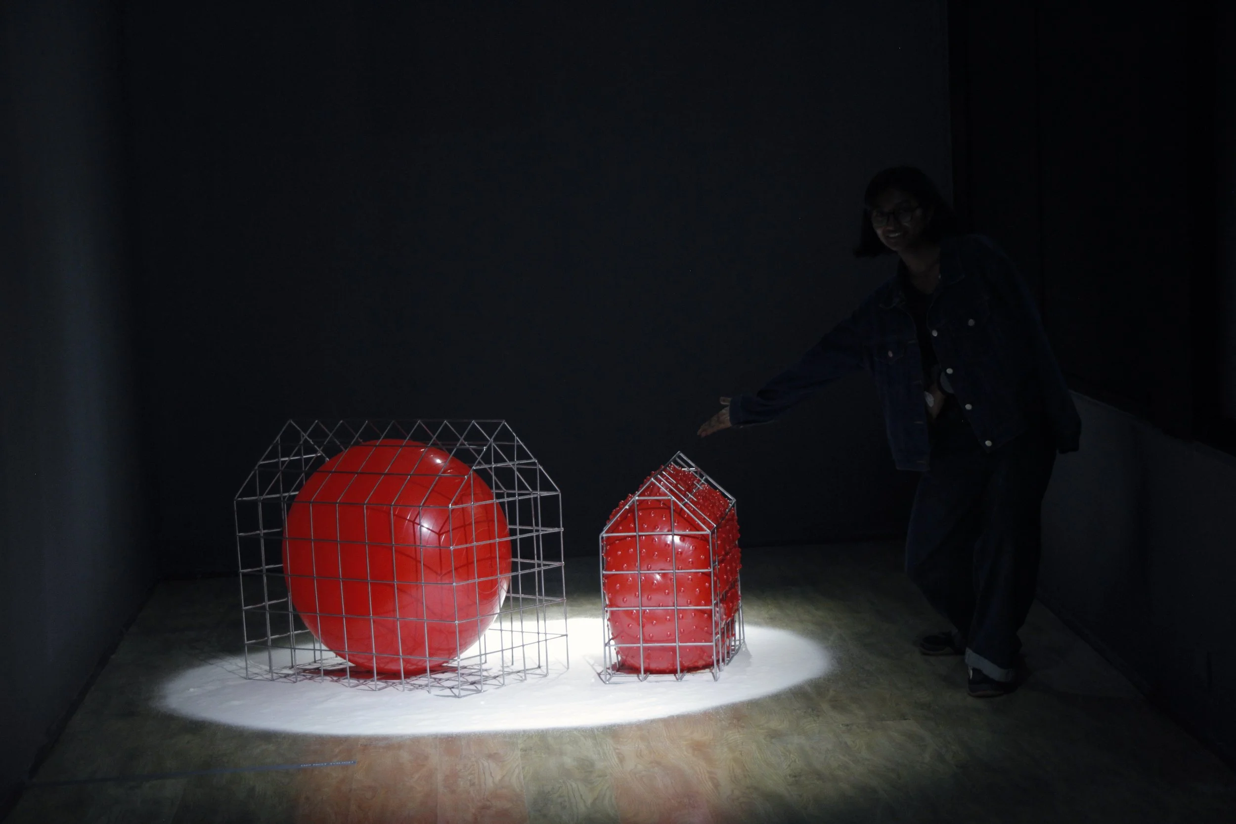 Dark exhibit with illuminated red geometric sculptures, including a large sphere, cube, and elongated shape, behind wireframe cages, with a person reaching towards the sculptures.
