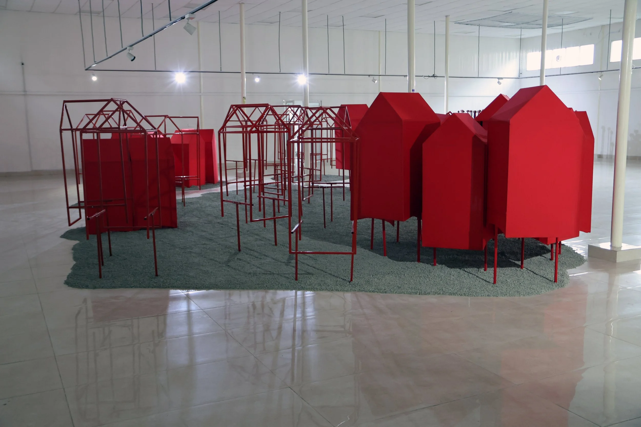 An art installation featuring red geometric structures on a green carpet inside a spacious room with white walls and large windows.