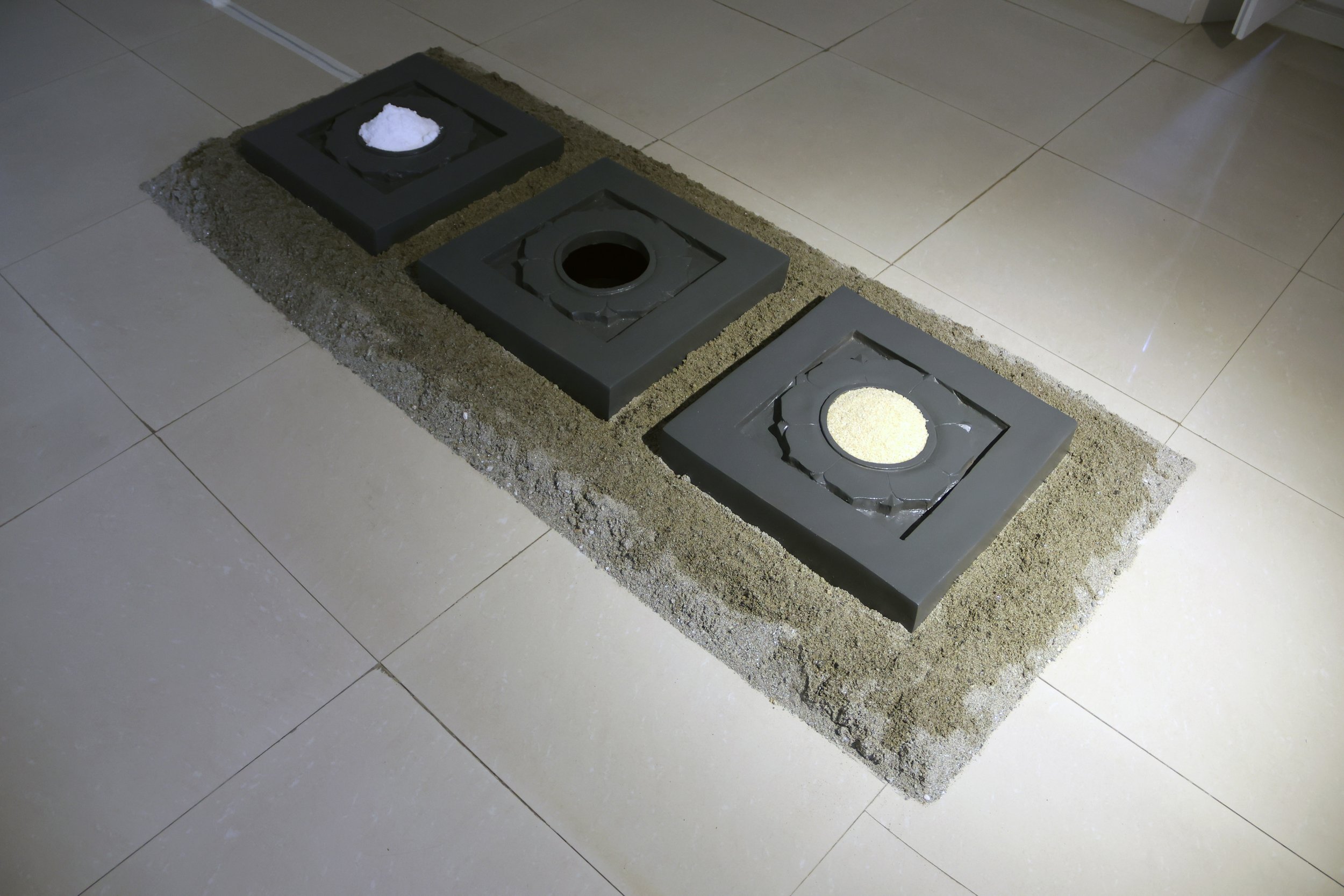 A construction site with three black square frames set in a bed of sand on a tiled floor, two of which contain white materials and one appears to be a hole.