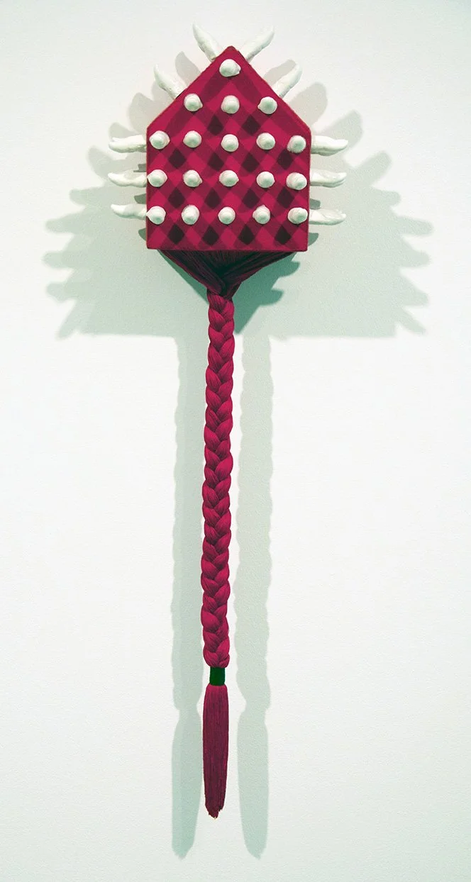 A decorative craft resembling a fantasy weapon or character with a red checkered body, white protrusions, a long braided tail, and shadowed on a white wall.