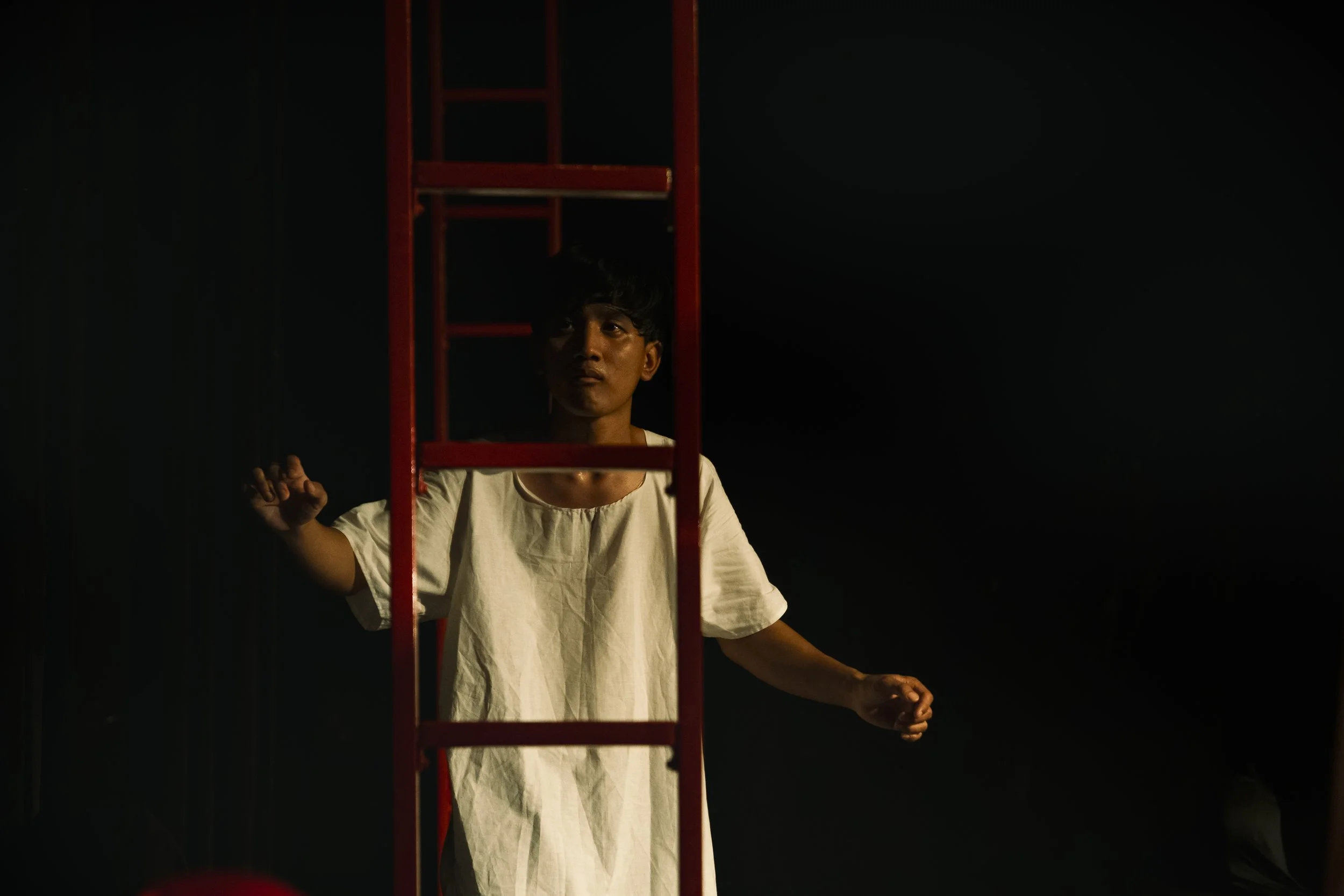 A performer in a white shirt stands behind a red ladder against a dark background on the stage.