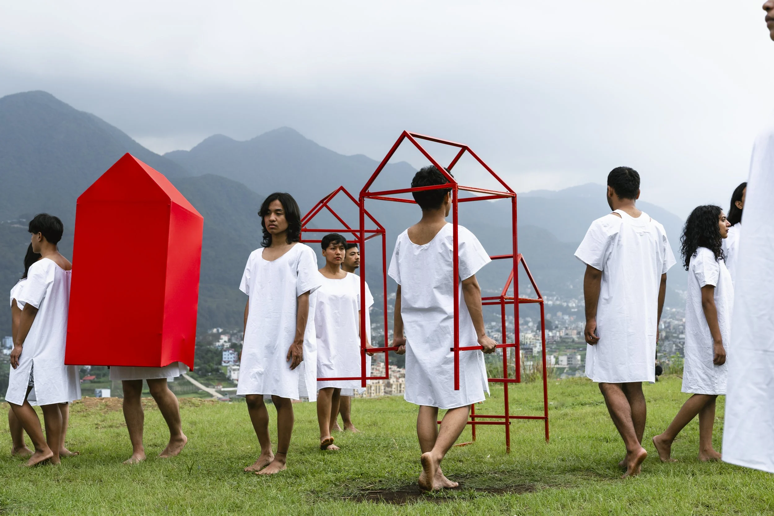 Group of people dressed in white robes standing outdoors on green grass, with a background of mountains and cloudy sky, some holding red geometric frames and a large red block.