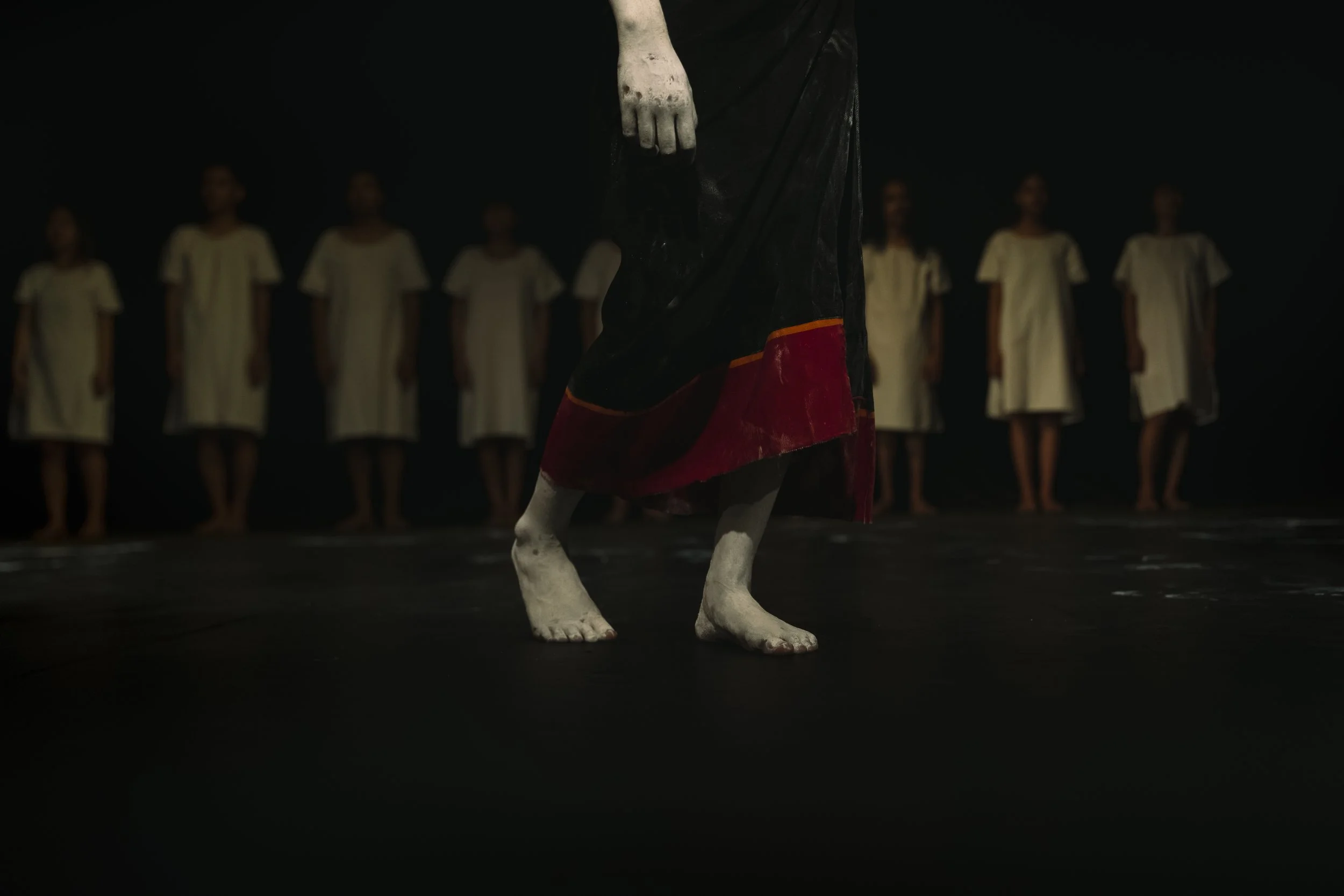 A scene from a theatrical performance showing a person dressed in black and red in front of a line of performers in white dresses. The background is dark.