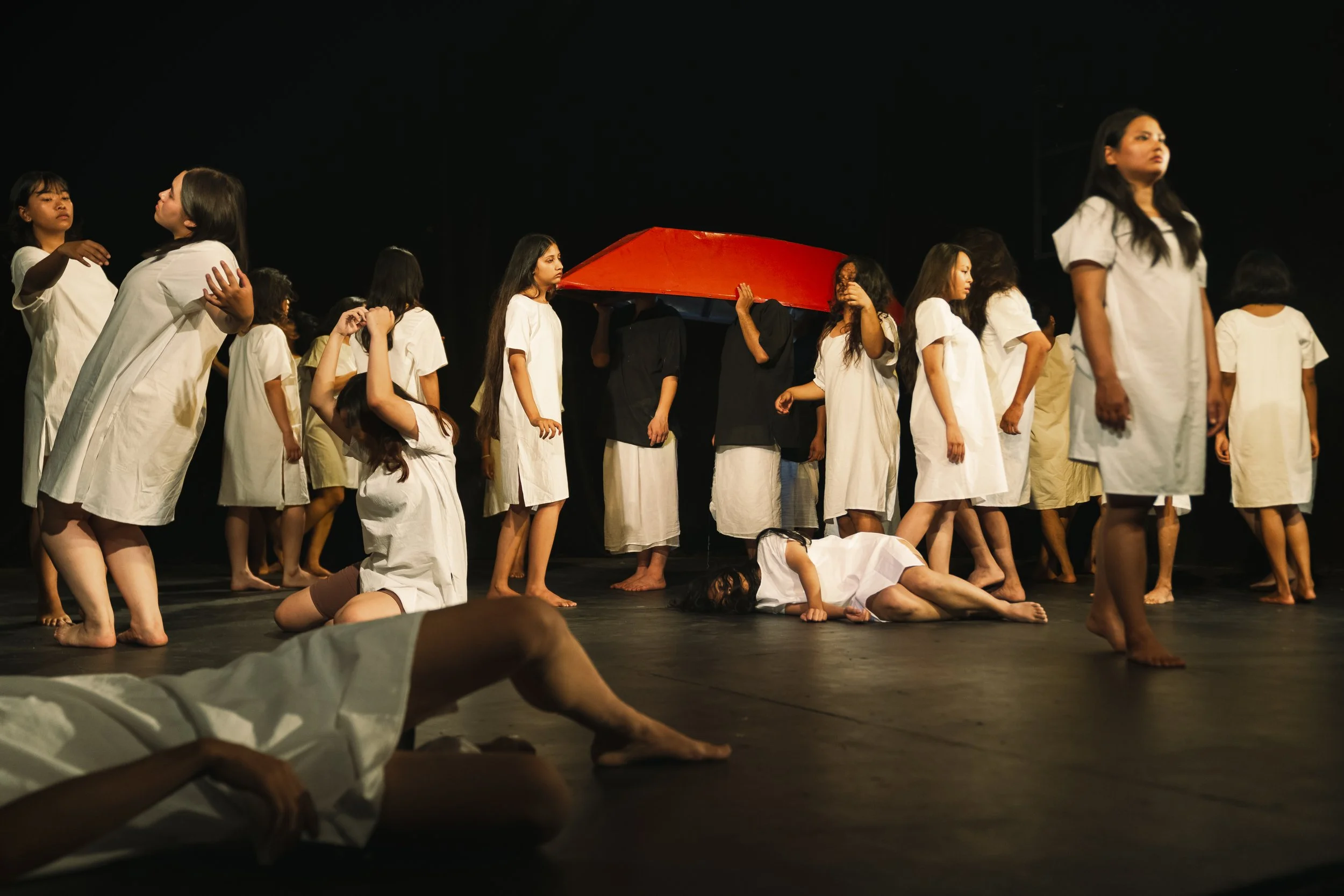 Performers in white and black clothing participate in a theatrical performance or dance on a stage, with some lying on the floor and others standing and holding a red object.