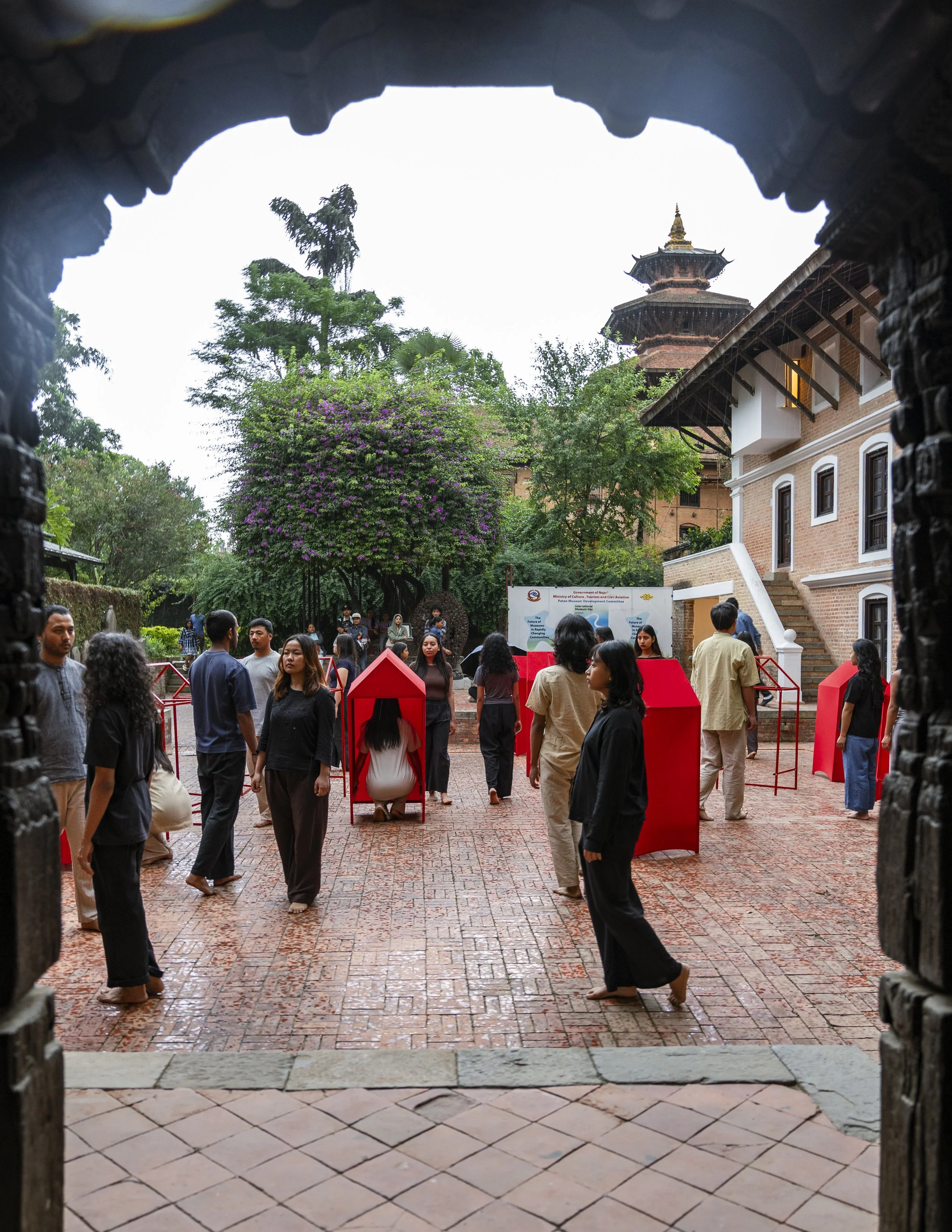 People gathered in a courtyard with traditional architecture, including a pagoda-style building, surrounded by trees and blooming shrubs, viewed from an arched entrance.