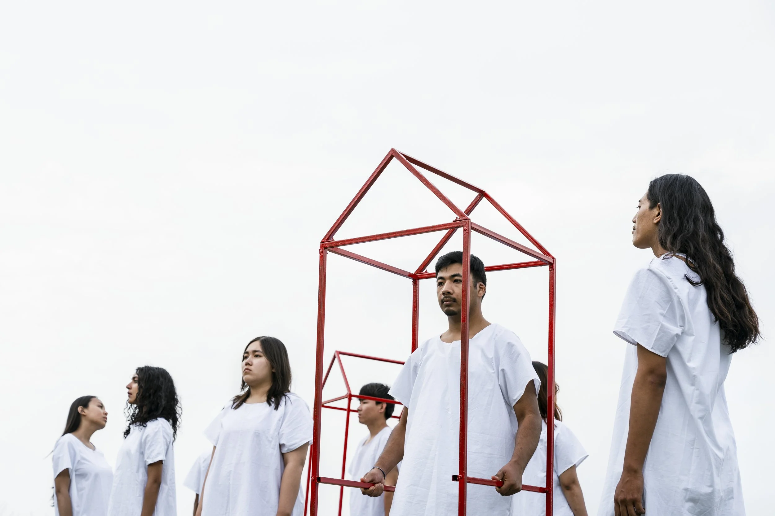 Group of young people standing outdoors, some holding red geometric frames, with a cloudy sky in the background.