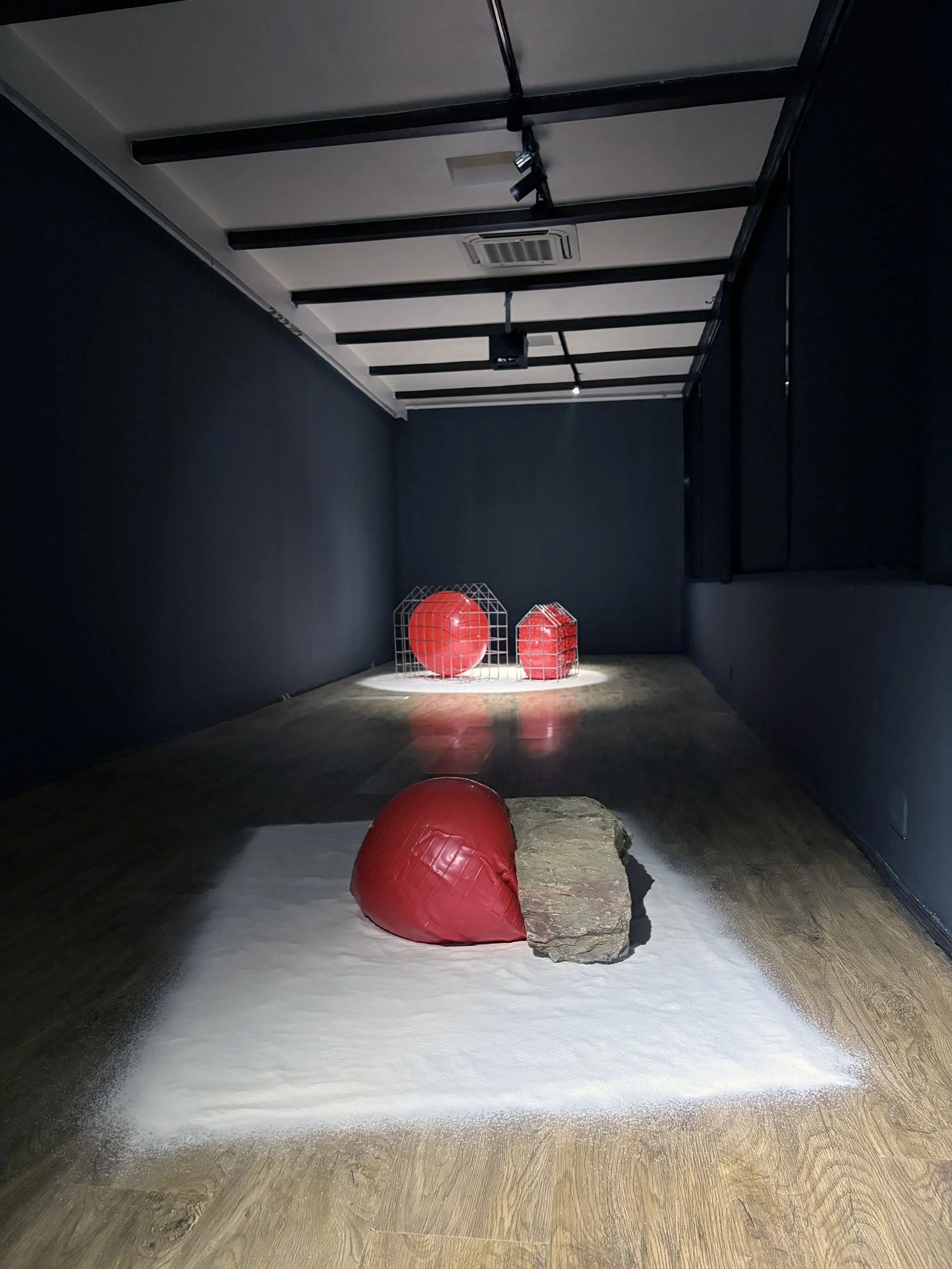 Art installation featuring red sports balls, a large rock, a white surface resembling snow, enclosed in a dark room with black walls and ceiling, and overhead lighting.