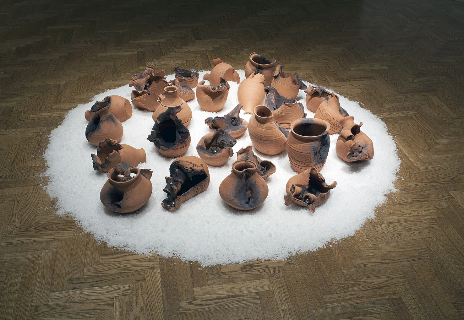 Collection of broken terracotta pots arranged on a white circular surface on a wooden floor in an art exhibit.